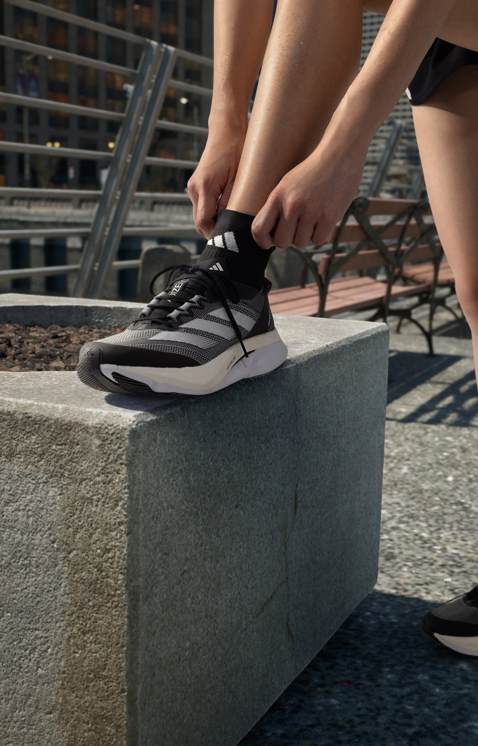 Person bindet die Schnürsenkel eines schwarzen und grauen Adidas Laufschuhs auf einer Betonbank im Freien.