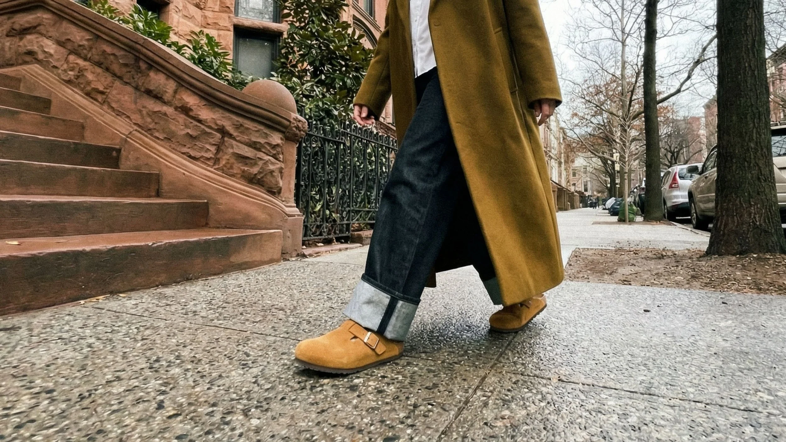 Nahaufnahme eines Spaziergängers, der eine braune Hose, braune Schuhe und einen langen braunen Mantel in einer städtischen Straße an einer Ecke trägt.