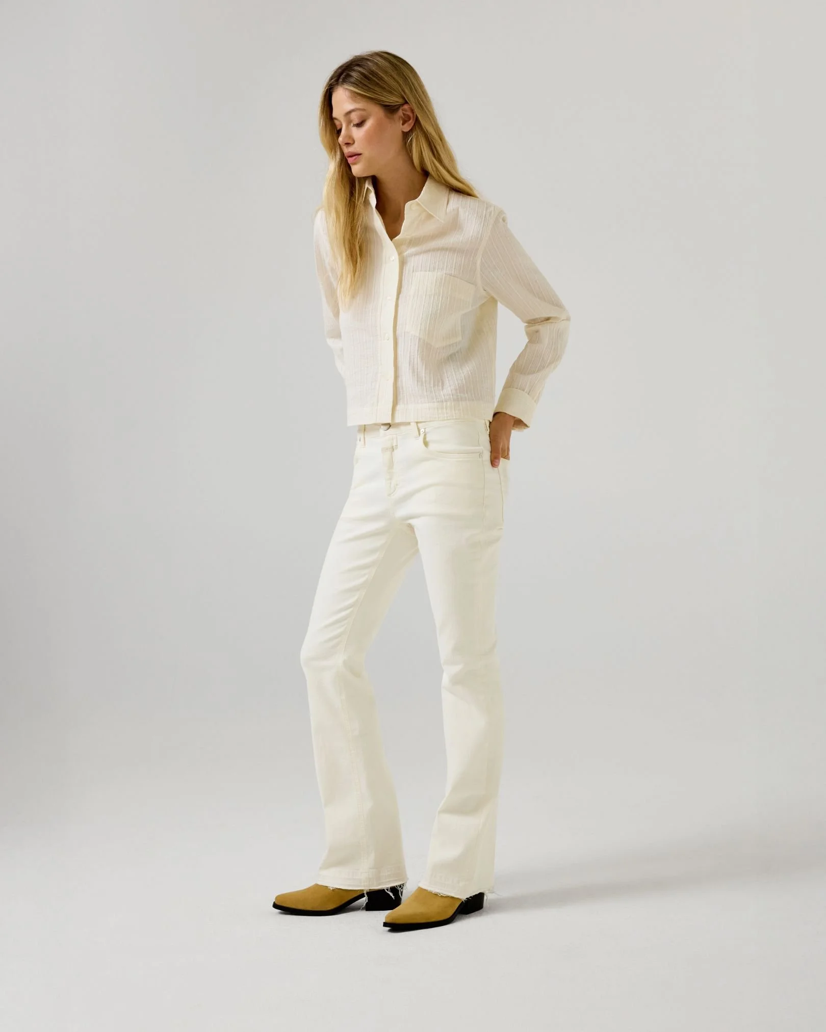 Frau trägt eine cremefarbene Bluse, weiße Jeans und braune Boots, steht vor einem hellgrauen Hintergrund.