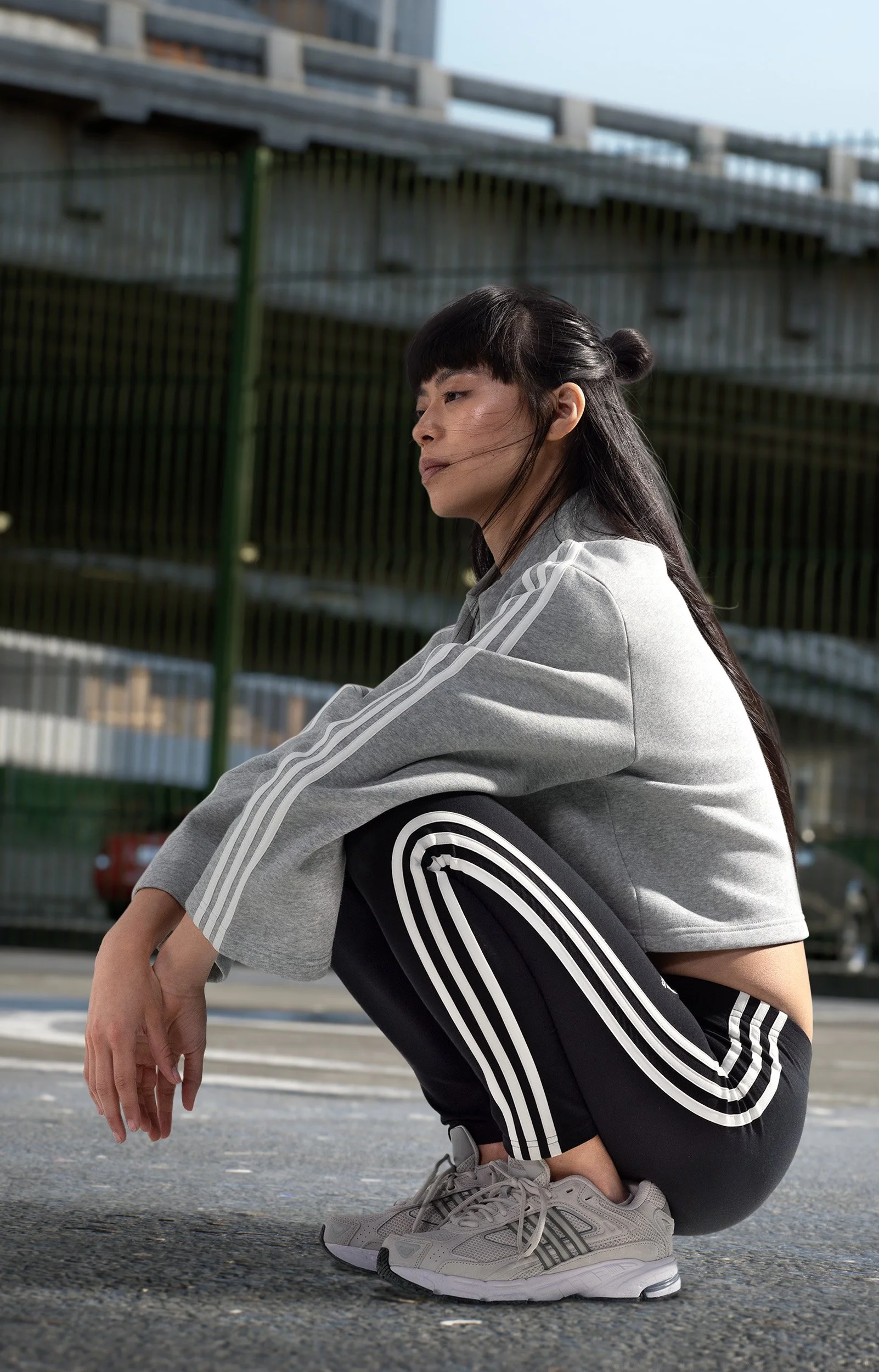 Junge Frau in Sportkleidung im squatting auf der Straße, umgeben von urbaner Umgebung mit Brücke und Gebäuden im Hintergrund.