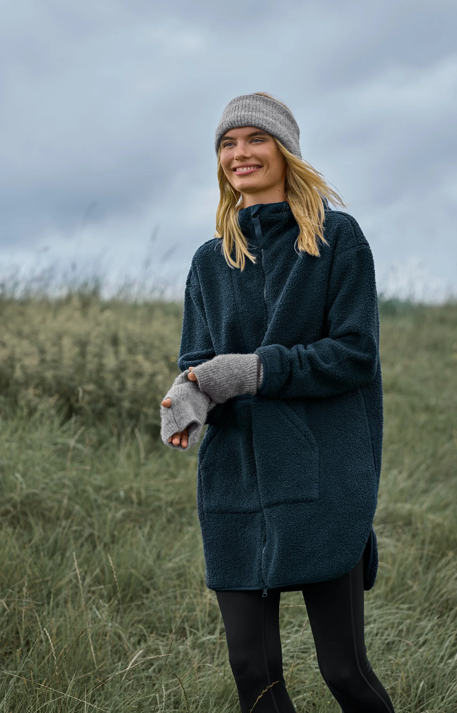 Eine junge Frau steht in einer grünen Wiese bei bewölktem Himmel, trägt einen grauen Stirnverband, eine dunkelblaue Outdoor-Jacke und graue Handschuhe.
