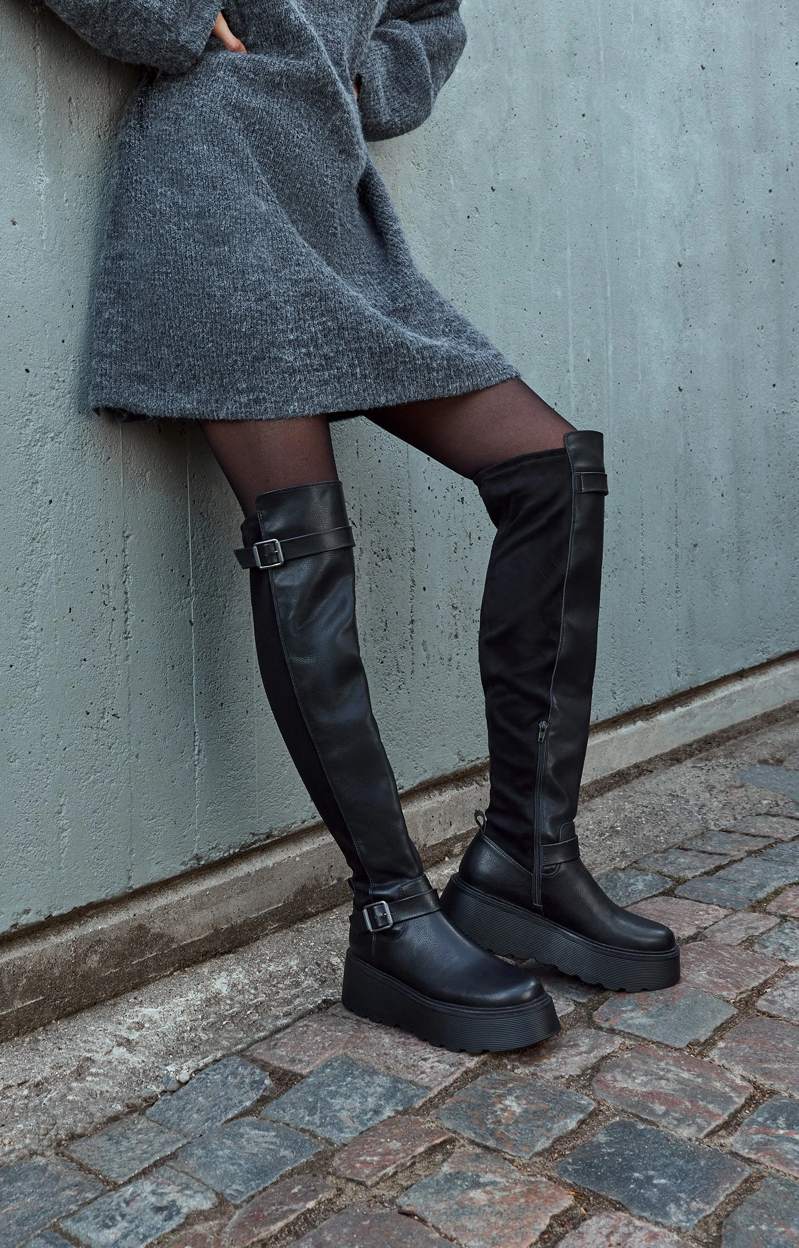 Frau trägt schwarze Overknee-Stiefel, dunkle Strumpfhose, grauen Mantel und steht an einer Mauer auf gepolstertem Boden.