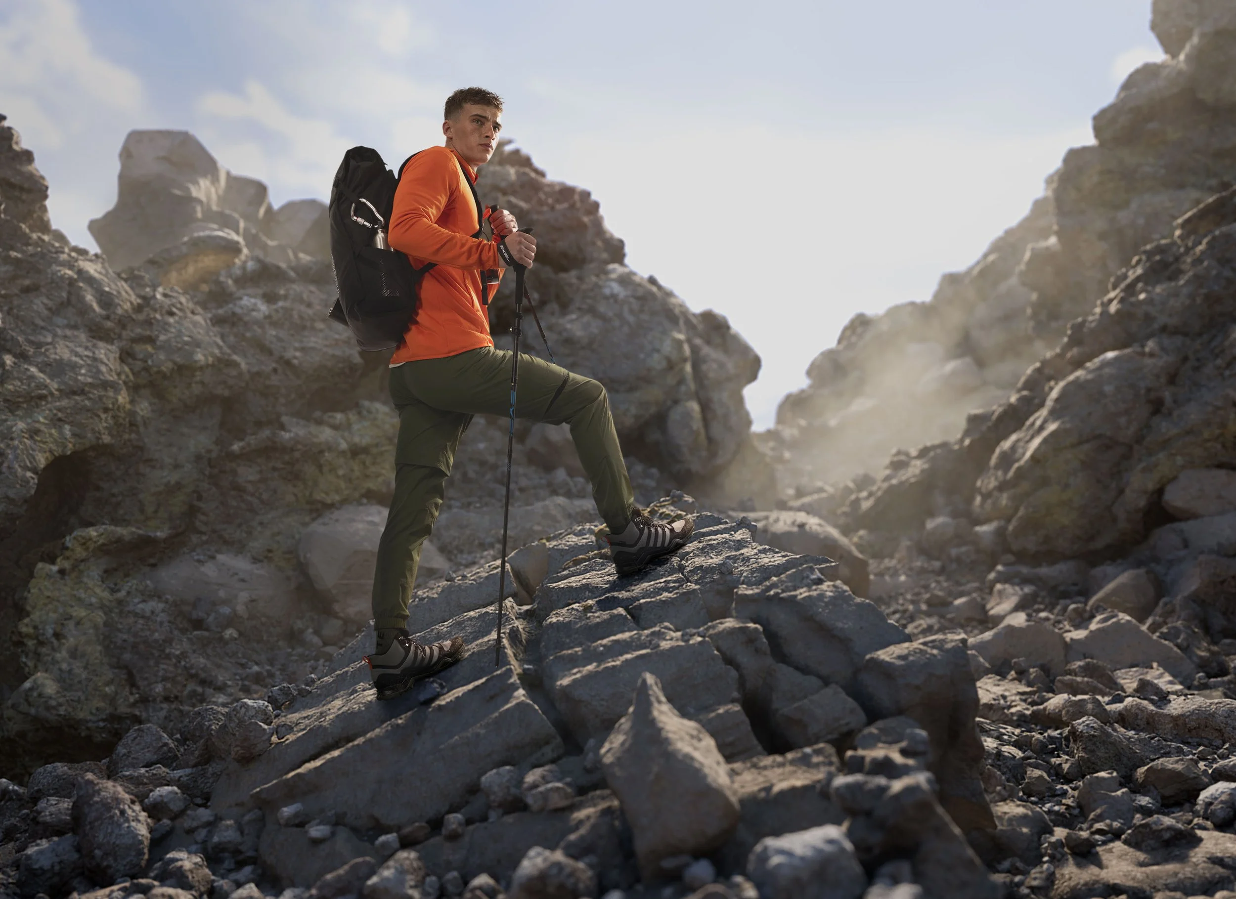 Mann beim Wandern in felsiger Gebirgsregion trägt orange Jacke und grüne Hose, benutzt Trekkingstöcke, trägt Rucksack, erklimmt einen steilen Felsen bei Sonnenlicht.