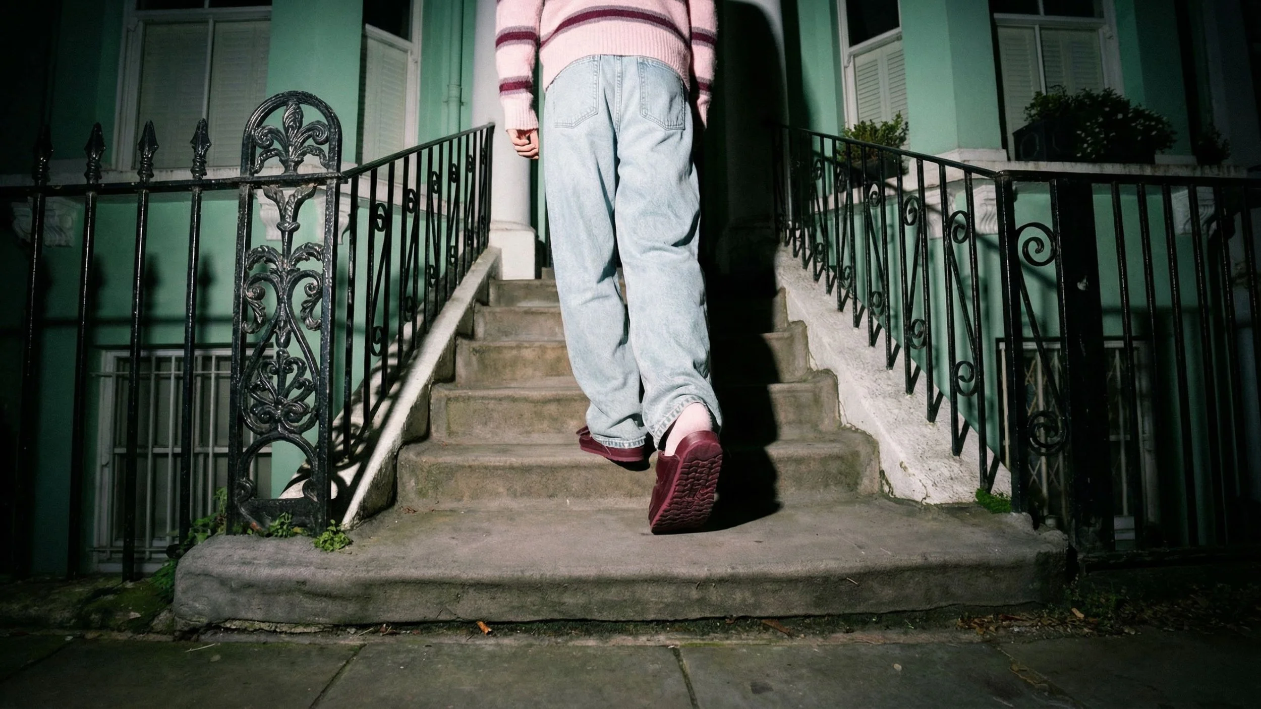 Blick auf eine Treppe, auf den grauen Stufen steht eine Person mit hellblauen Jeans und roten Schuhen, im Hintergrund ein grünes Haus mit schwarzen Geländern.