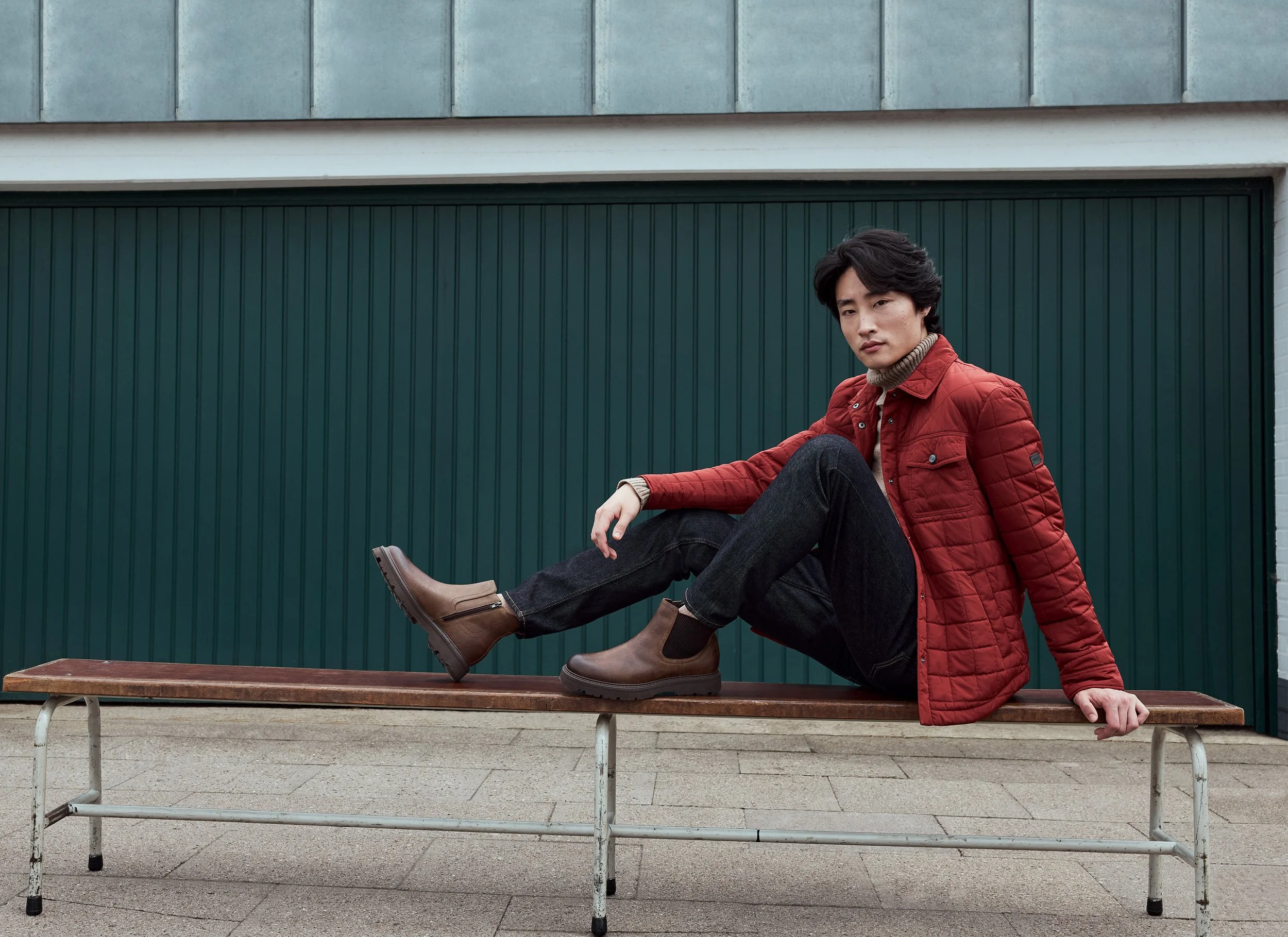 Ein junger Mann sitzt auf einer Bank vor einer grünen Garage, trägt eine rote Jacke, schwarze Hose und braune Stiefel, schaut in die Kamera.
