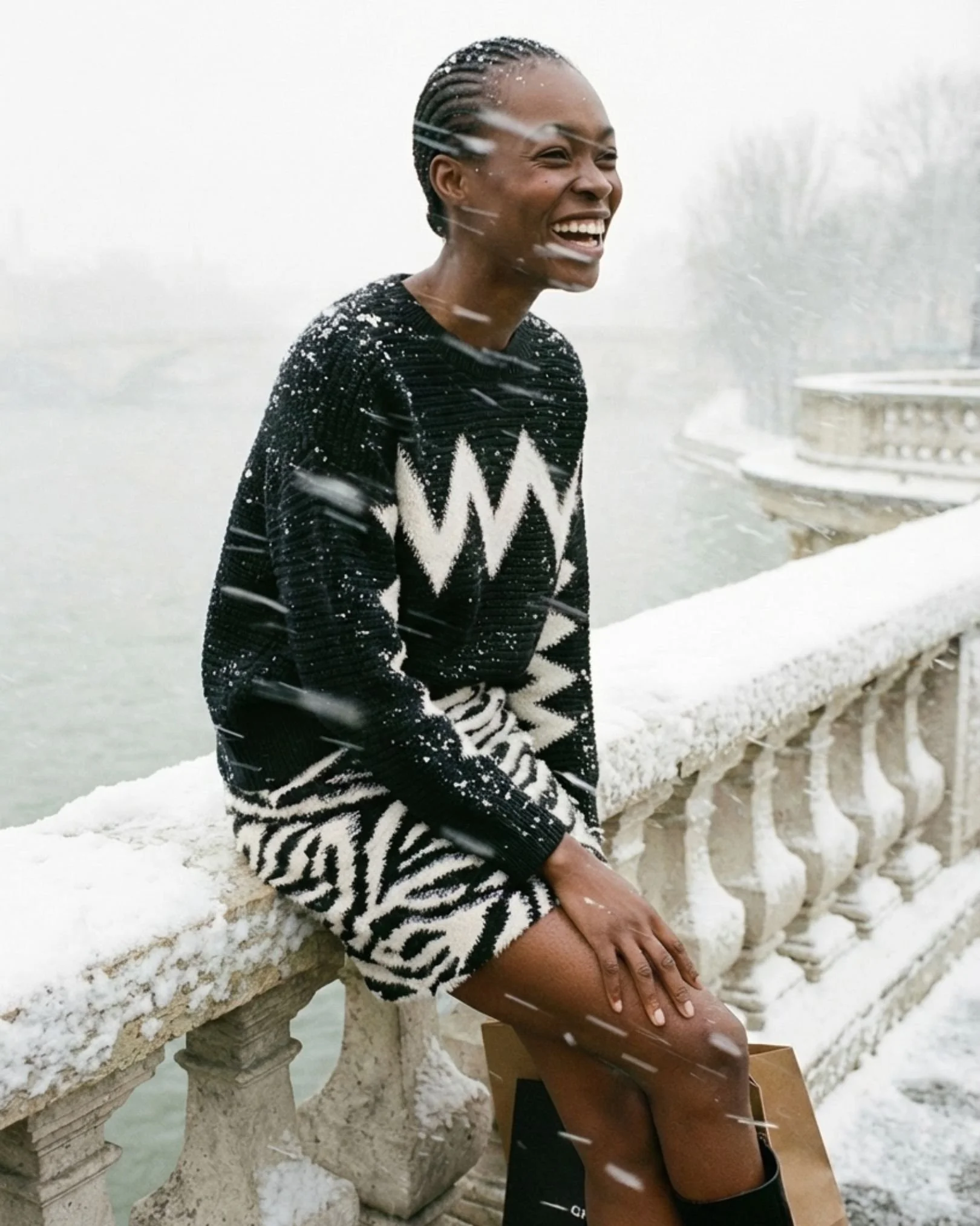 Eine lachende Frau mit Zopf, die im Schneesturm auf einer Brücke sitzt, trägt eine schwarze und weiße Wolljacke und ein Zebra-Muster Kleid.