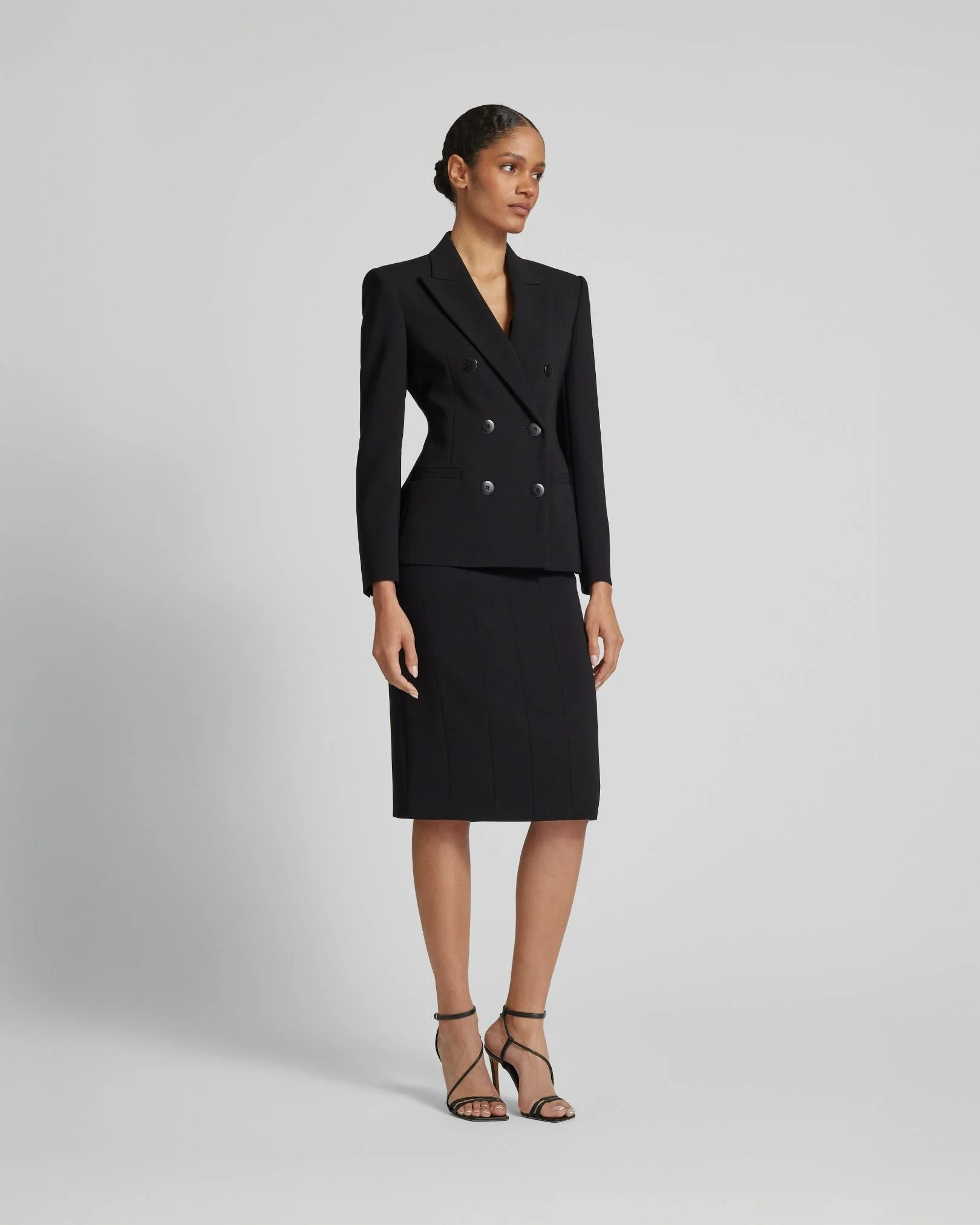 Frau in schwarzem Businesskostüm mit hochhackigen Sandalen vor grauem Hintergrund