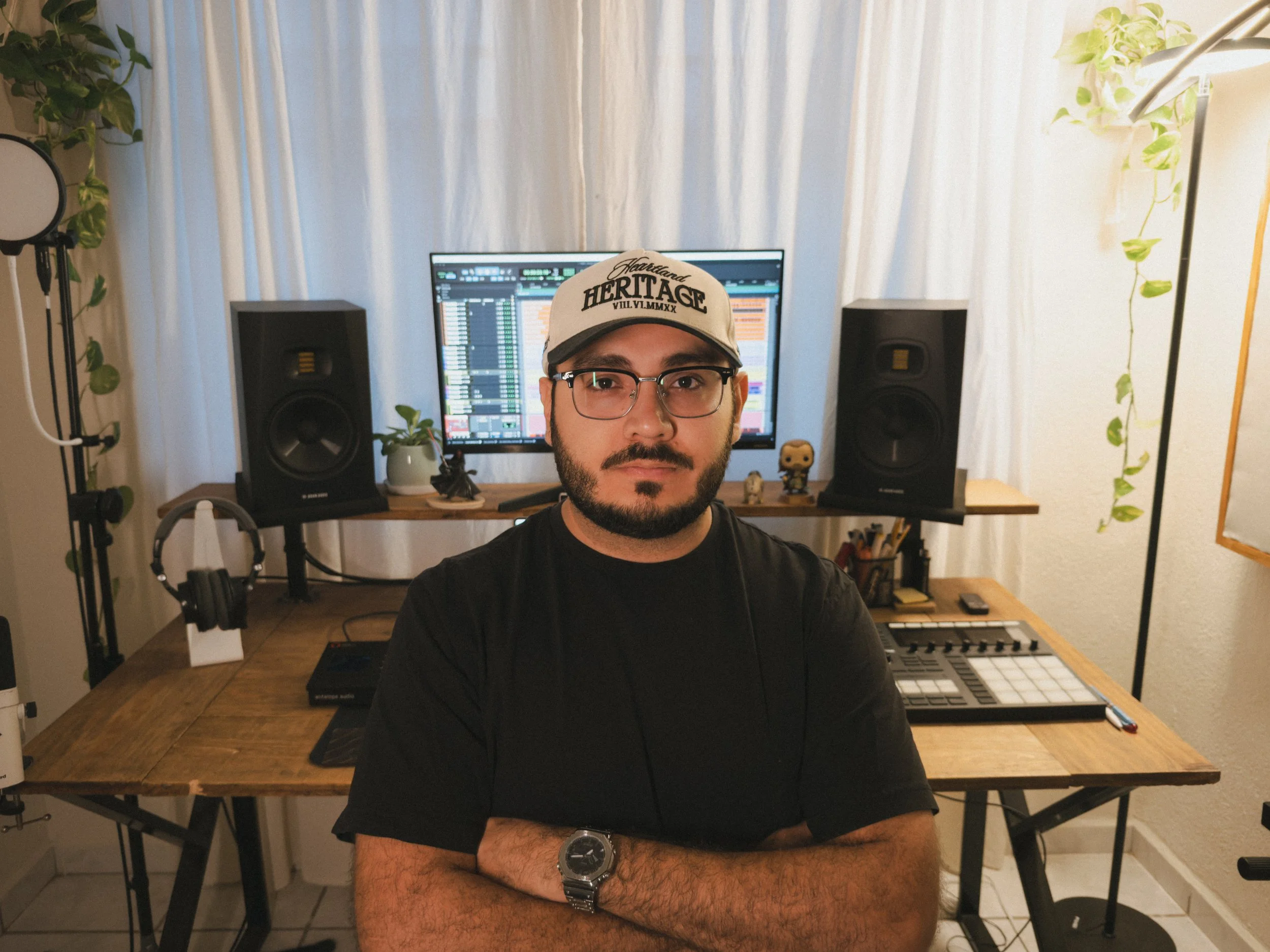 A man with glasses and a beard wearing a cap and black t-shirt stands with arms crossed in a home music studio with a computer, speakers, and music equipment.