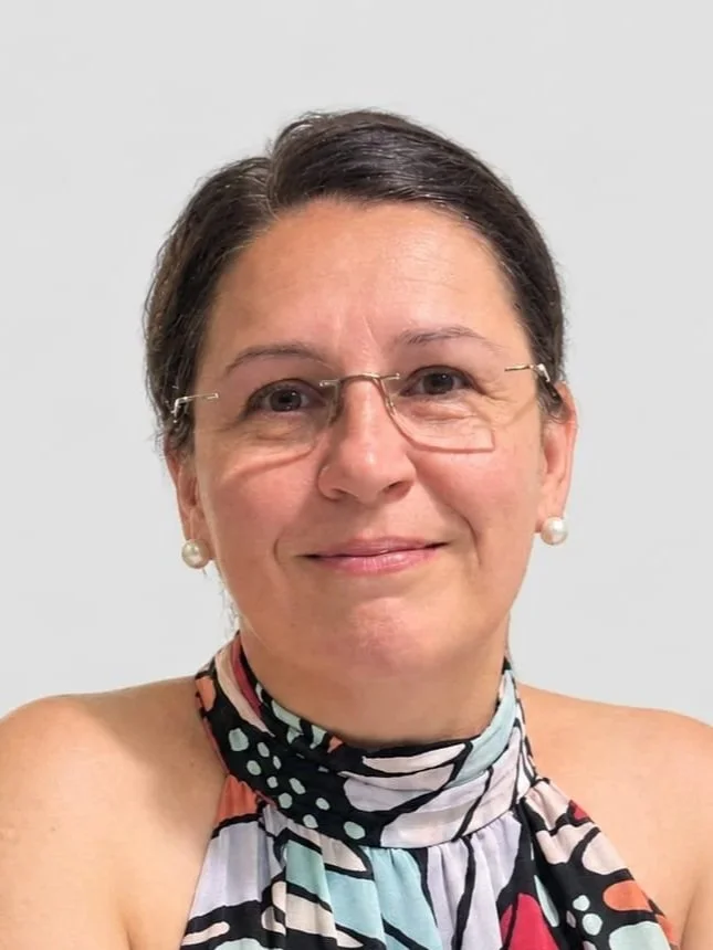 Eine Frau mit kurzen dunklen Haaren, Brille und Perlenohrringen, die lächelnd in die Kamera schaut, trägt ein farbenfrohes, gestreiftes Halstuch ohne Schultern sichtbar.