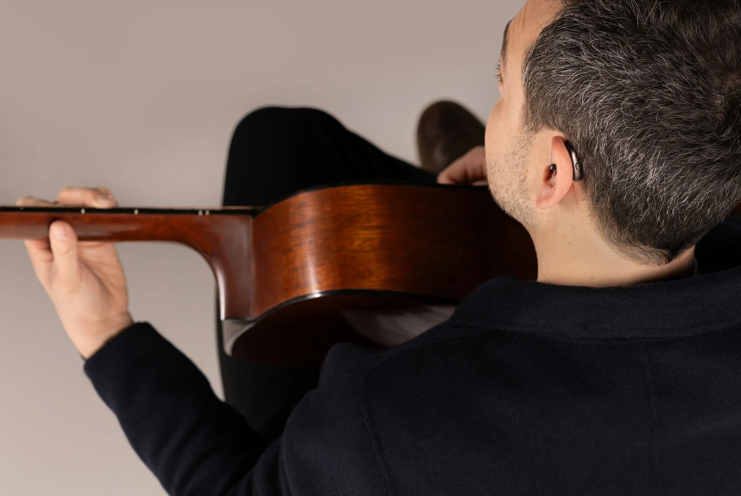 Close-up of a man playing an acoustic guitar, viewed from behind and slightly to the side, with short gray hair and wearing a black shirt.