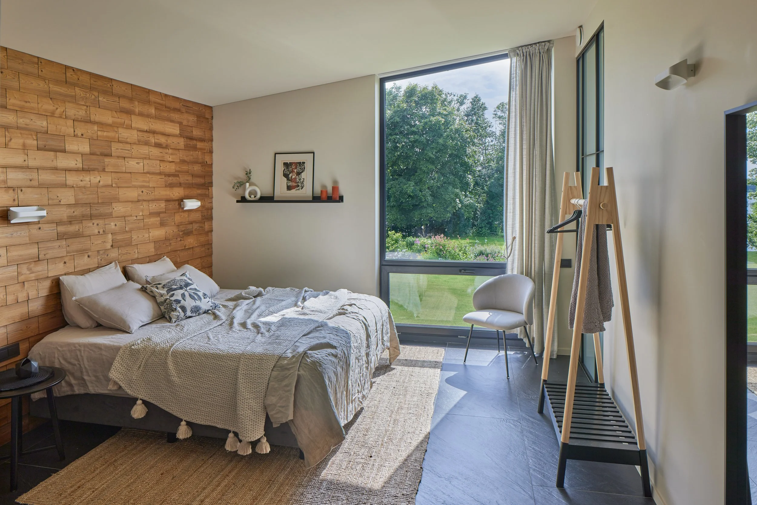 Modern bedroom with a wooden accent wall, white bedding, large window with green outdoor view, chair, and coat rack.