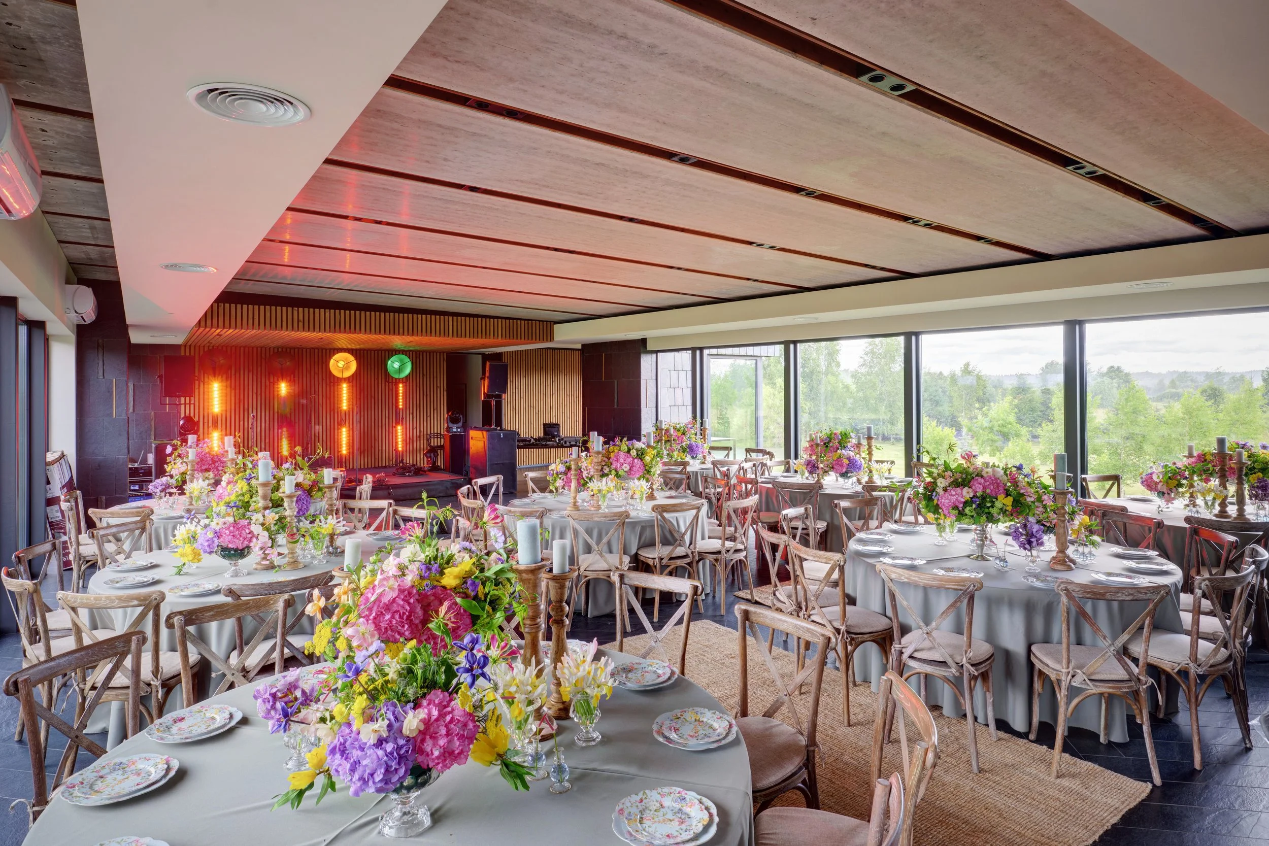 A decorated banquet hall with round tables covered in white tablecloths, adorned with floral centerpieces, candles, and floral plates. There are wooden chairs around each table. In the background, a stage with colorful lighting and speakers is visible, and large windows on the right side reveal a lush green outdoor landscape.