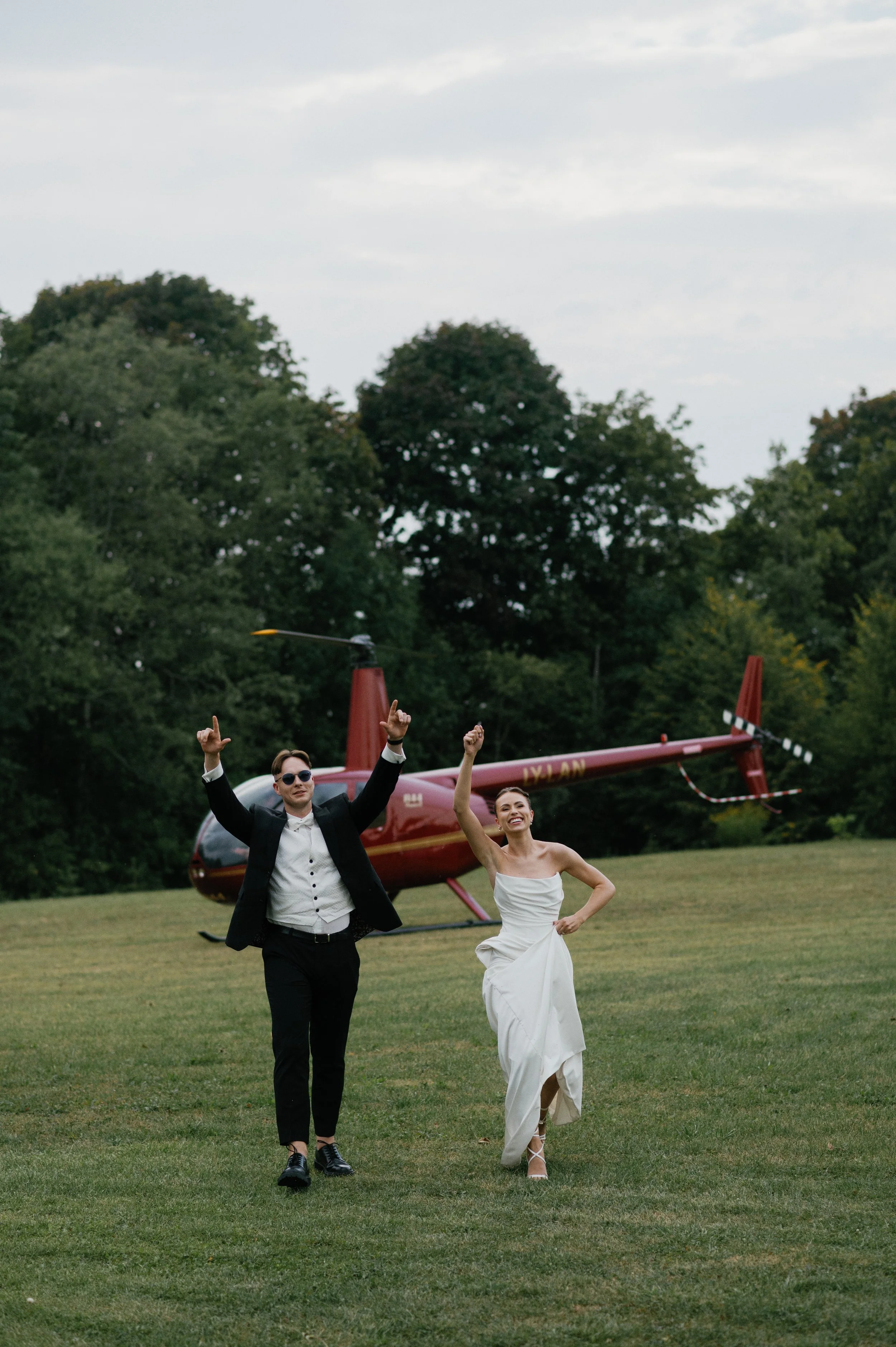A joyful couple in wedding attire running on a grassy field in front of a red helicopter, with trees and a cloudy sky in the background.
