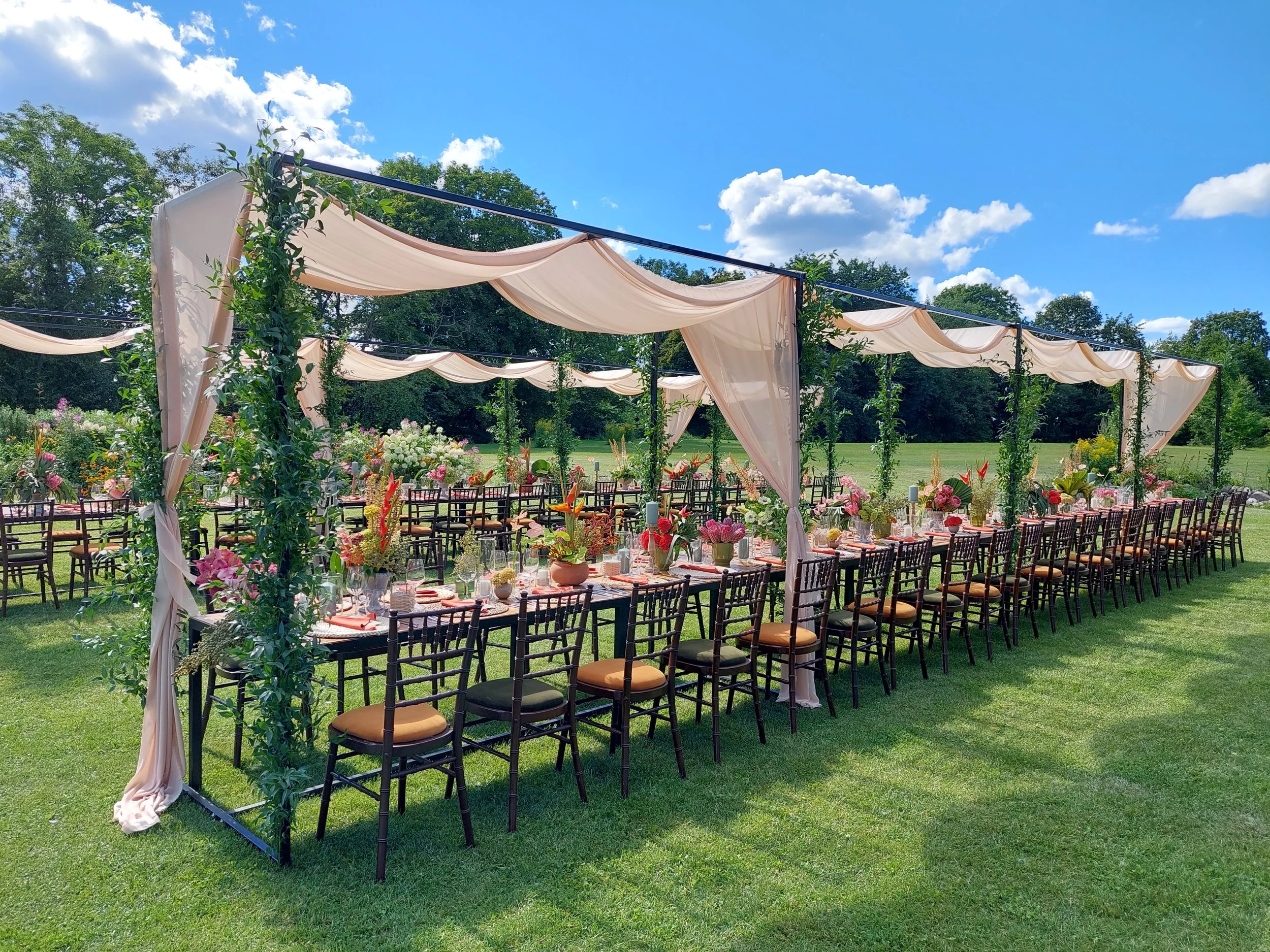 Outdoor wedding reception table setup in a lush green field under a blue sky with fluffy white clouds, featuring long tables with floral arrangements, candles, and surrounded by dark wooden chairs with brown cushions.