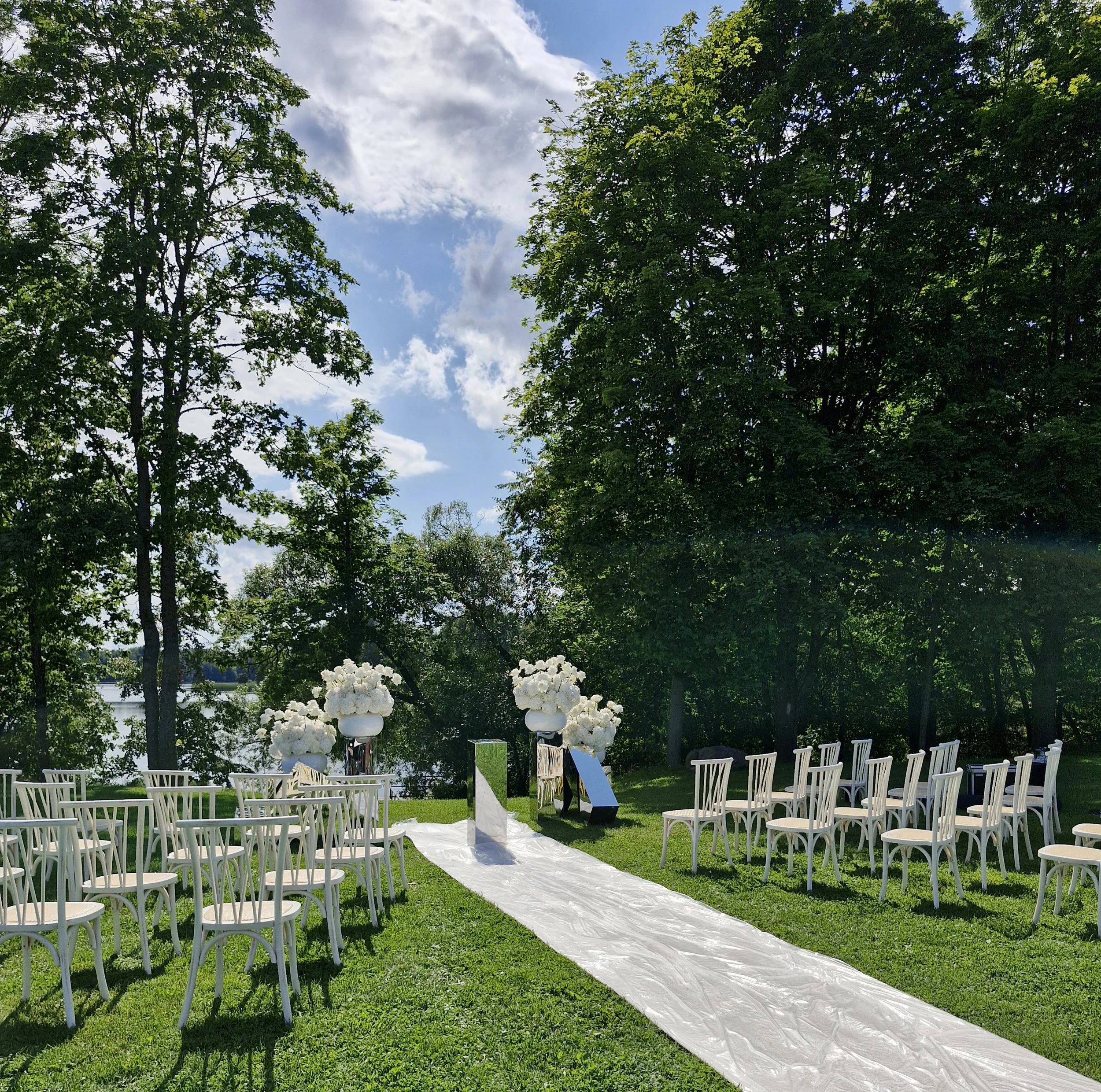 Outdoor wedding ceremony setup with white chairs on either side of a white aisle runner, decorated with white floral arrangements, green trees, a water body in the background, and a partly cloudy sky.