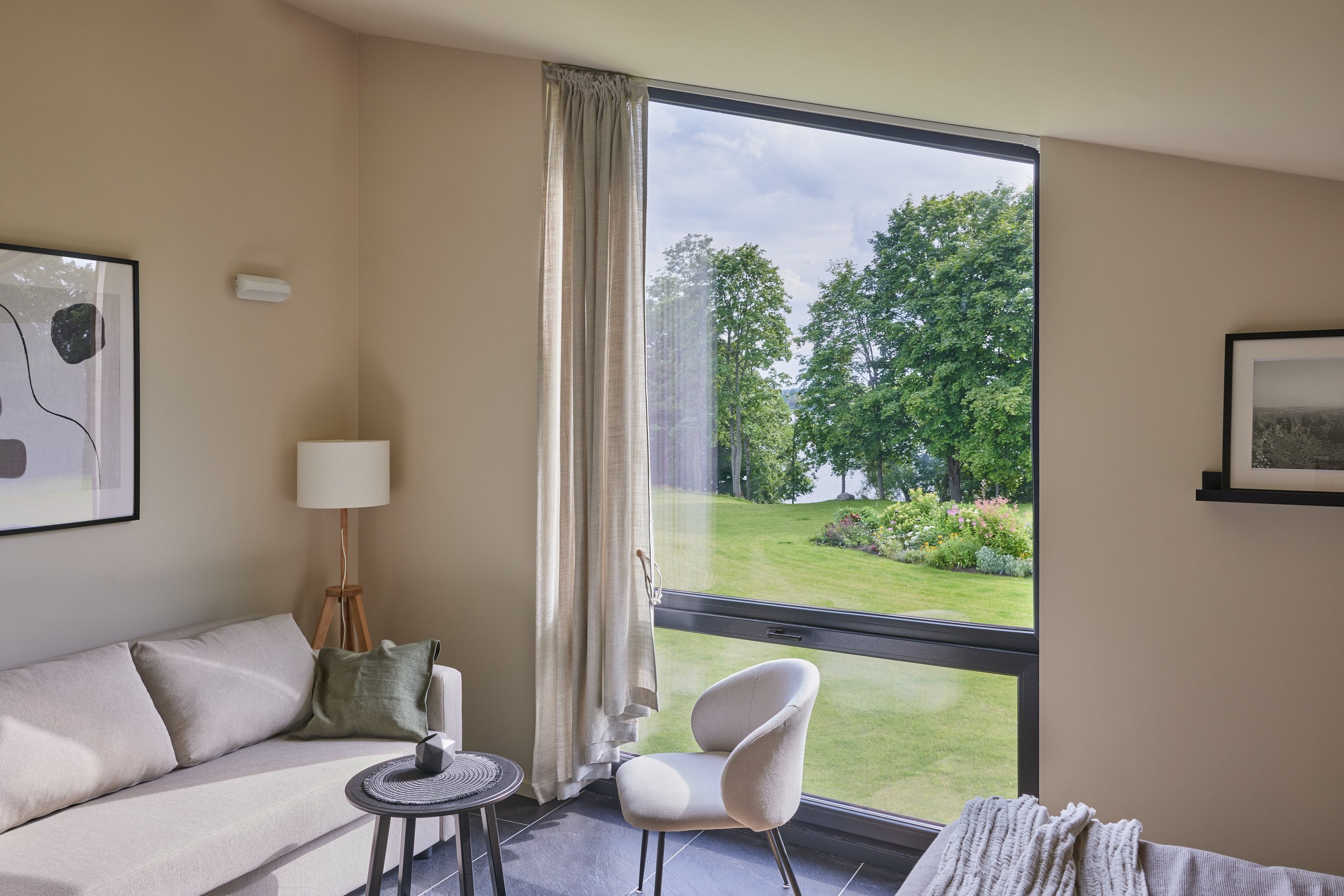 Living room with beige sofa, white armchair, black side table, lamp, and large window showcasing green trees and a garden outside.