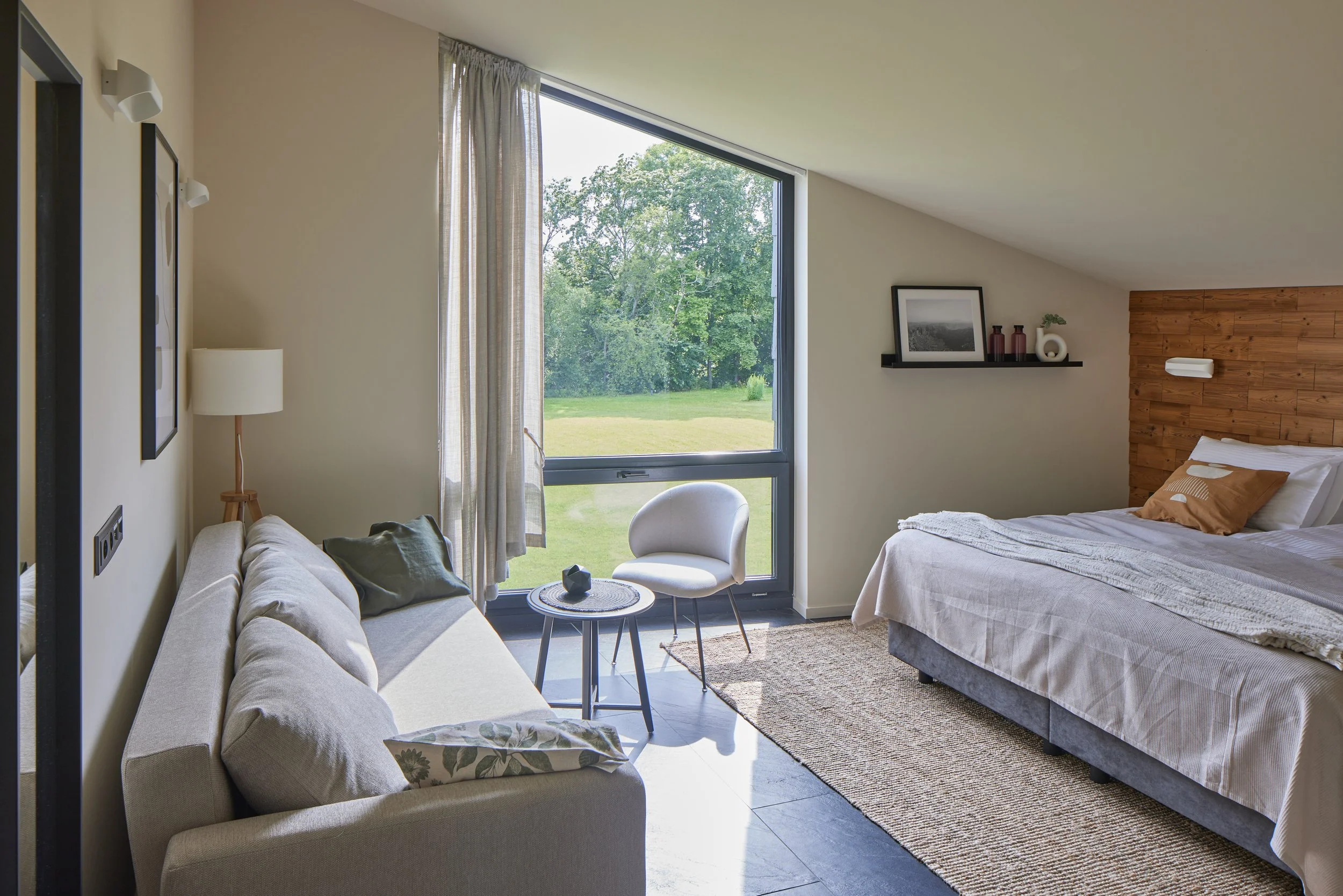 Bright bedroom with bed, beige sofa, round chairs, window with curtains, and outdoor greenery view.