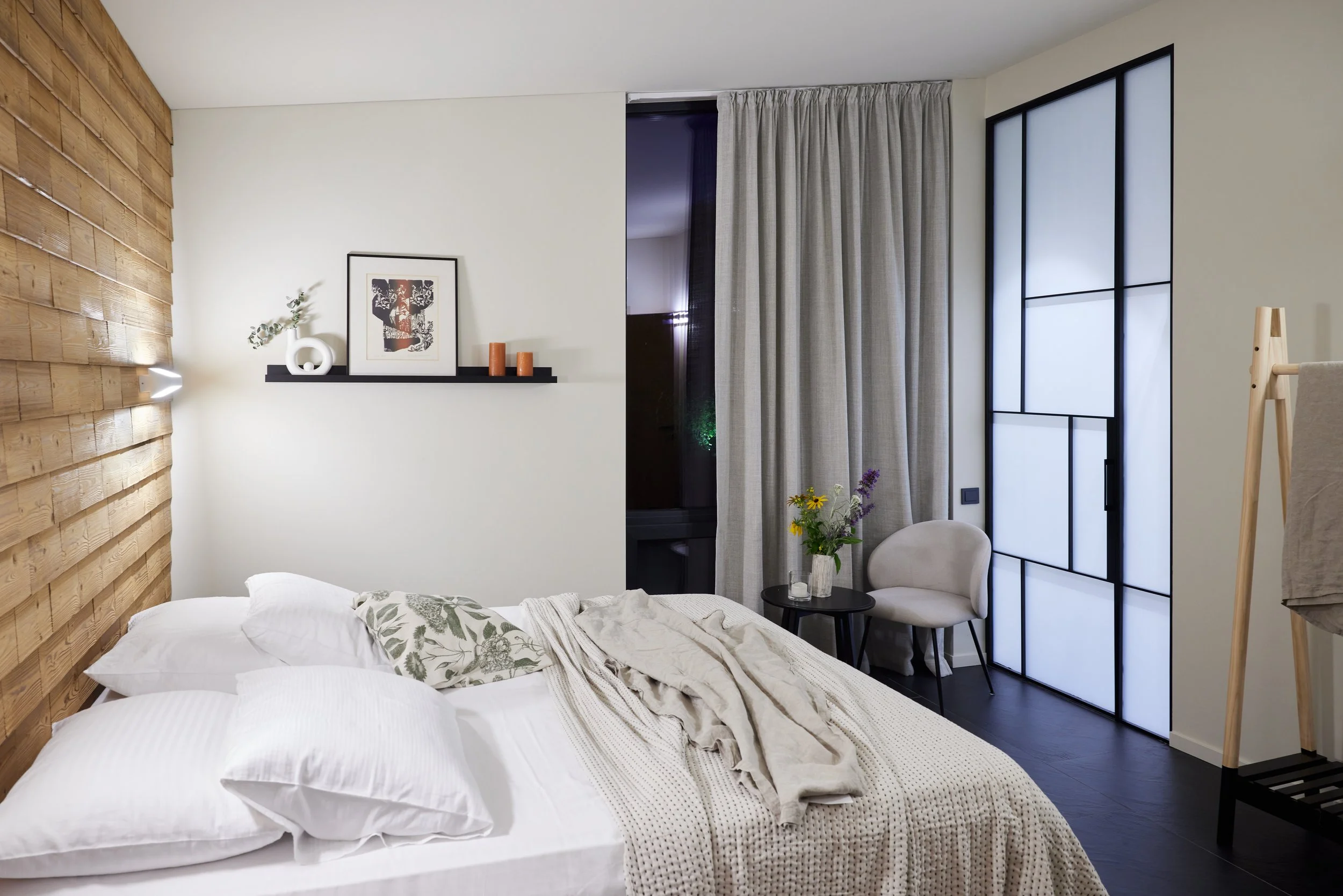 A modern bedroom with a white bed, floral pillows, and a beige blanket. Wooden accent wall on the left, a black shelf with decor and candles, beige curtains, and a black-framed sliding door. Small black side table with a vase of flowers and a cream chair near the window.