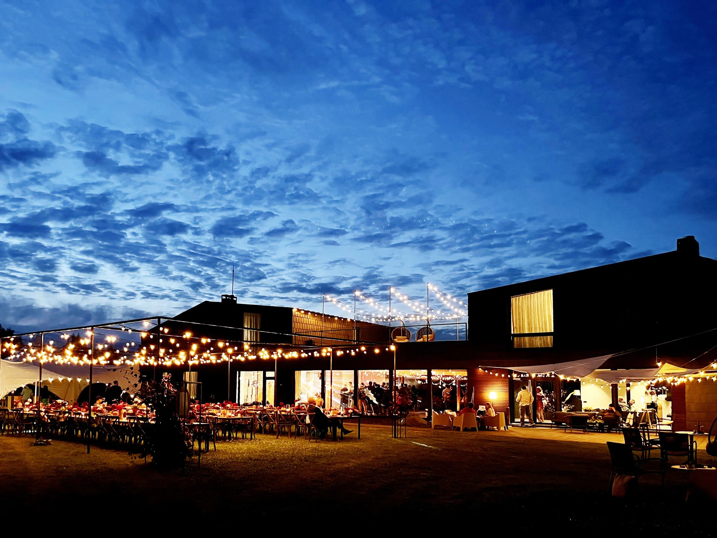 Outdoor evening gathering with string lights and people dining at tables under a partly cloudy sky.