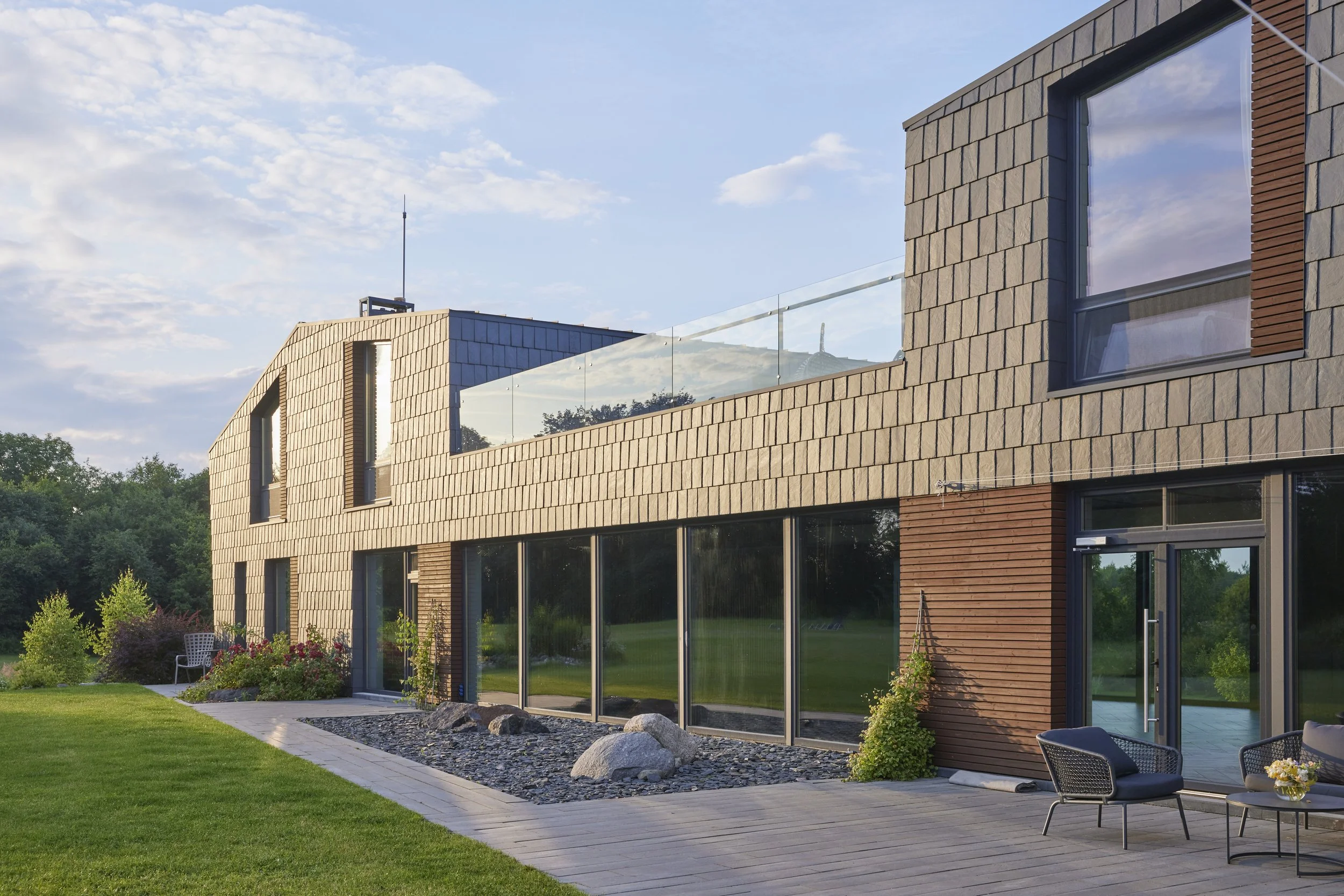 Modern multi-story house with large glass windows, wooden exterior, and a flat roof, surrounded by greenery and a lawn, under a partly cloudy sky.
