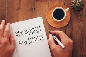 Open notebook with the words "New Mindset, New Results" written inside, held by a person's hands, on a wooden table with a cup of coffee on a small plate and a cactus plant nearby.