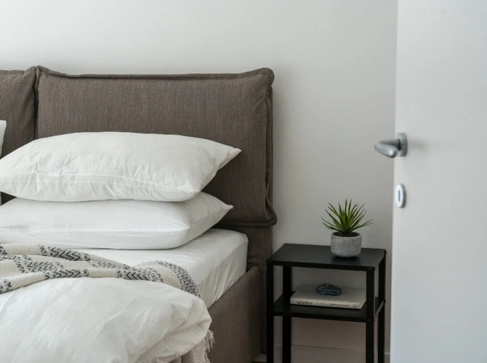 A cozy bedroom with a bed featuring a tufted brown headboard, white pillows, and a blanket. There is a black bedside table with a small potted plant and books.