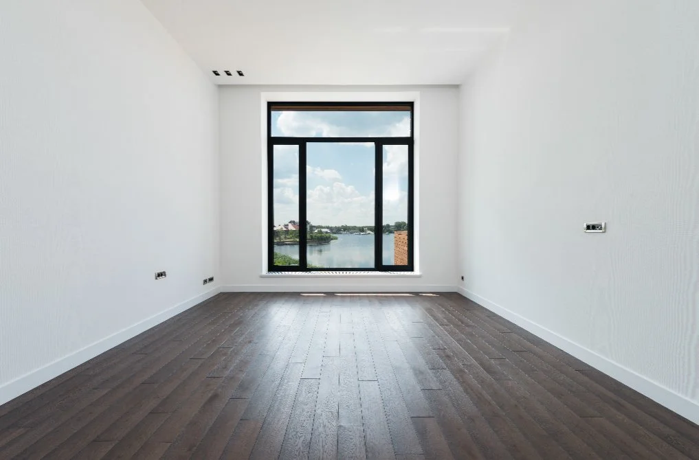 Empty room with white walls, dark wooden floor, and large window showing a view of a river, trees, and partly cloudy sky.