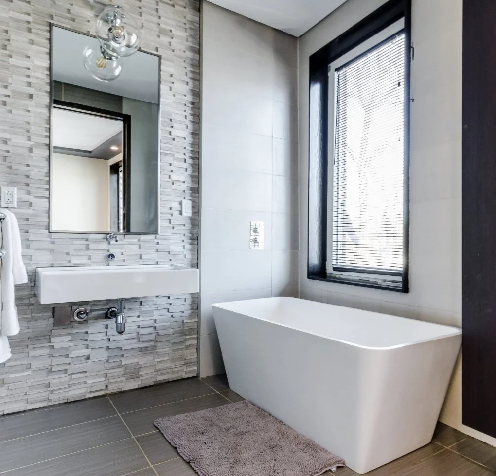 Modern bathroom with a white bathtub, a large window with blinds, a rectangular mirror over a small sink, and a textured gray wall.