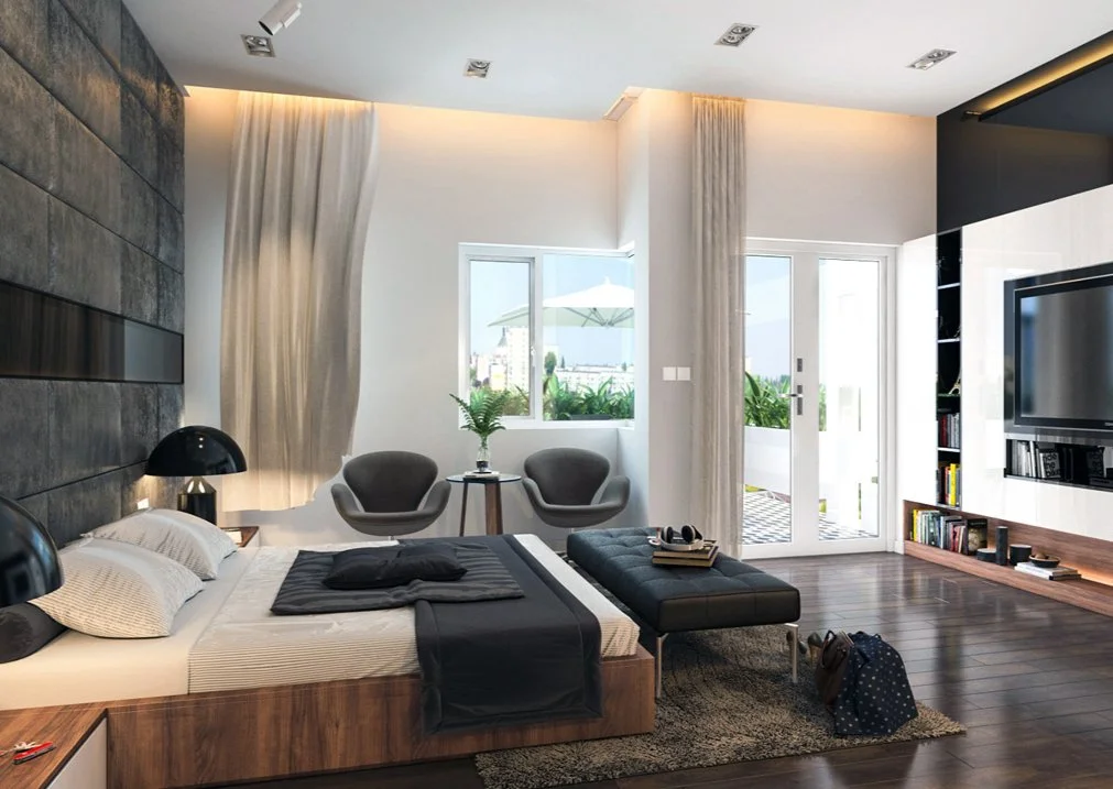 Modern bedroom with a large bed, sitting area with two chairs and a small table, sliding glass door opening to a balcony with outdoor furniture, television on the wall, and a bookshelf.