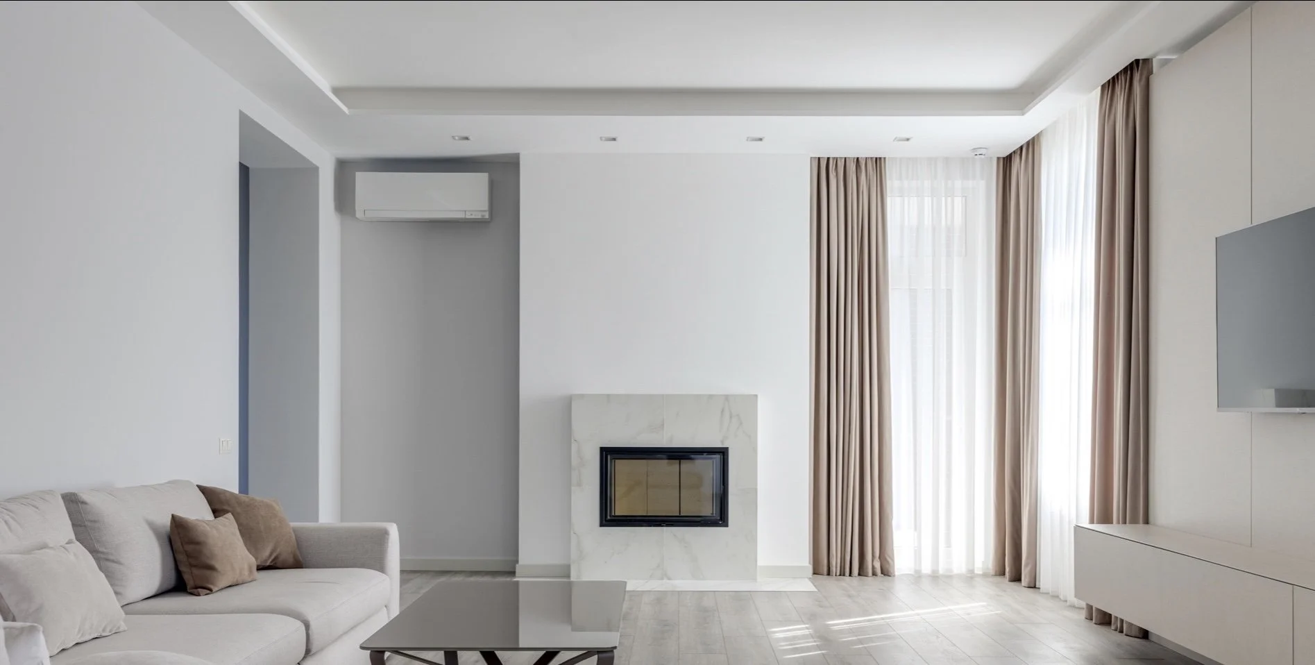 Modern living room with white walls, beige sofa with cushions, a white marble fireplace, large windows with beige curtains, and a mounted flat-screen TV.