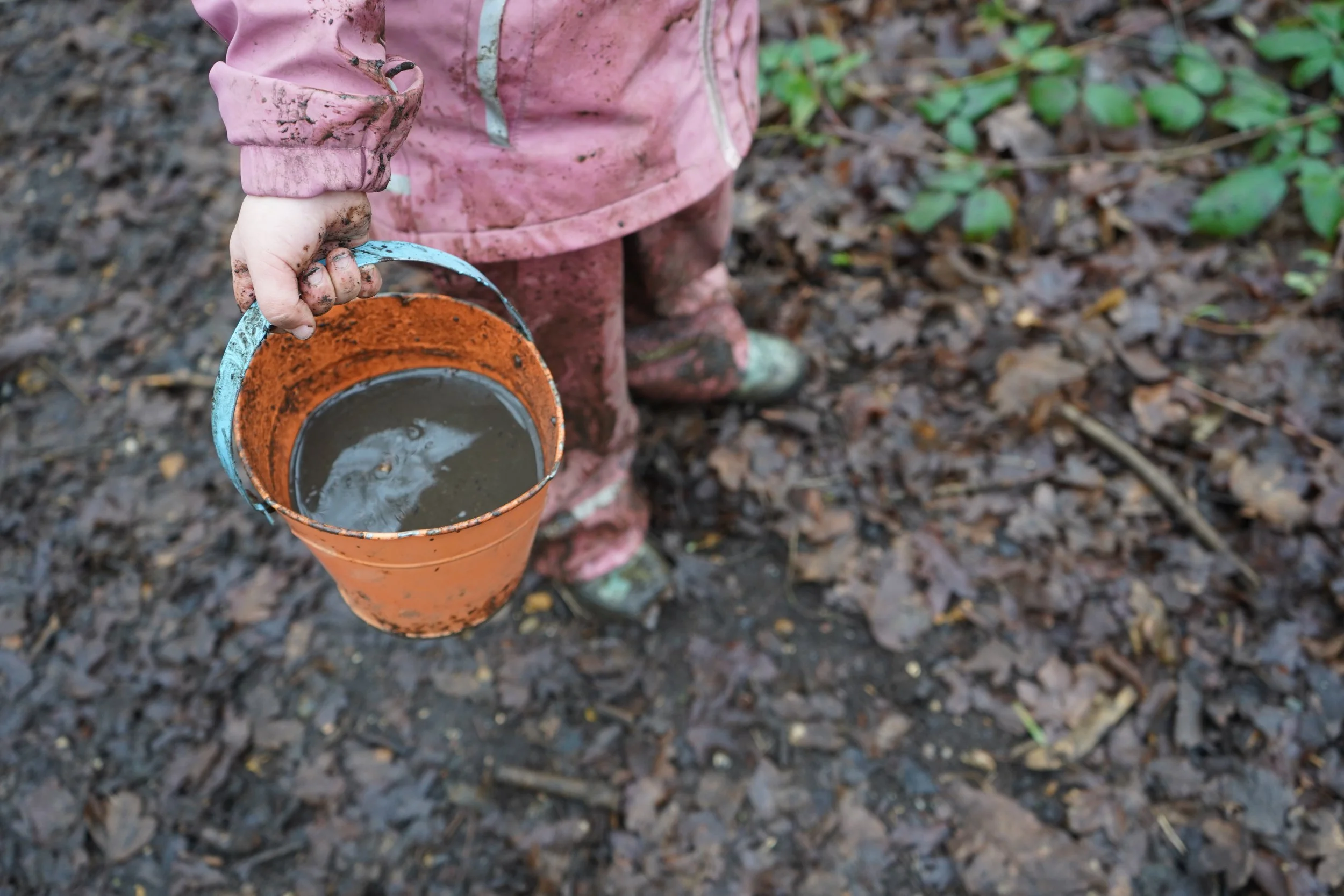Child in pink raincoat holding orange bucket filled with muddy water, standing on leaf-strewn ground.