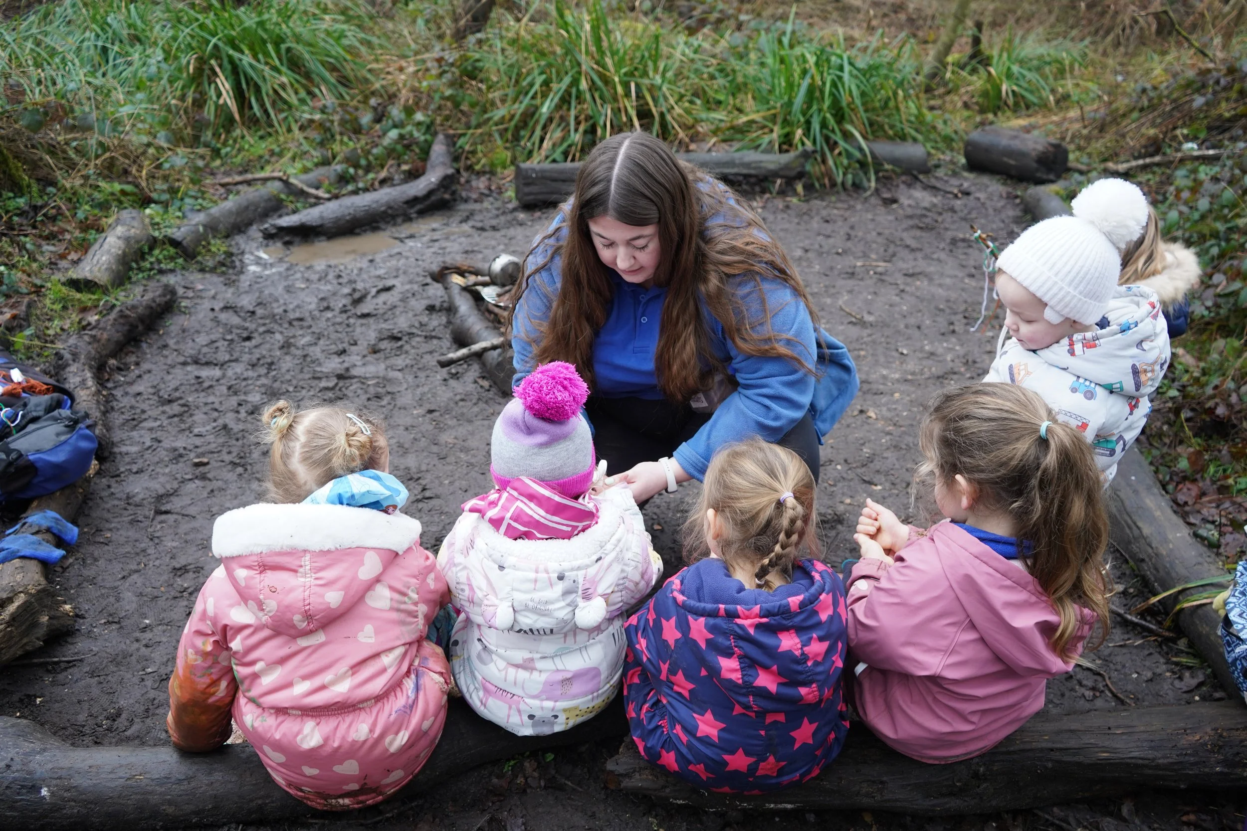 A nursery manager assists a group of children sitting on logs in the forest. Children are dressed in winter clothing, engaged in an activity, surrounded by nature with green plants in the background.