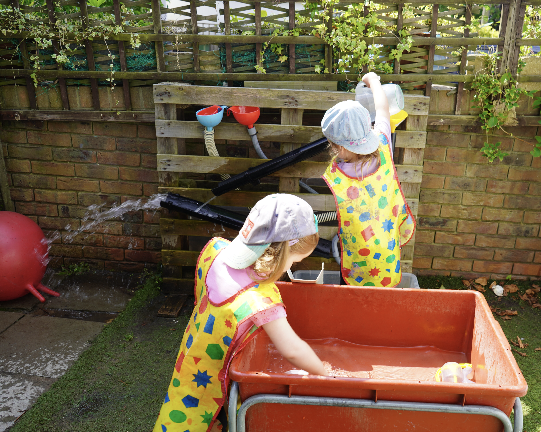 Children playing with water in an outdoor activity setup with colorful aprons and caps, using funnels and pipes against a wooden pallet.