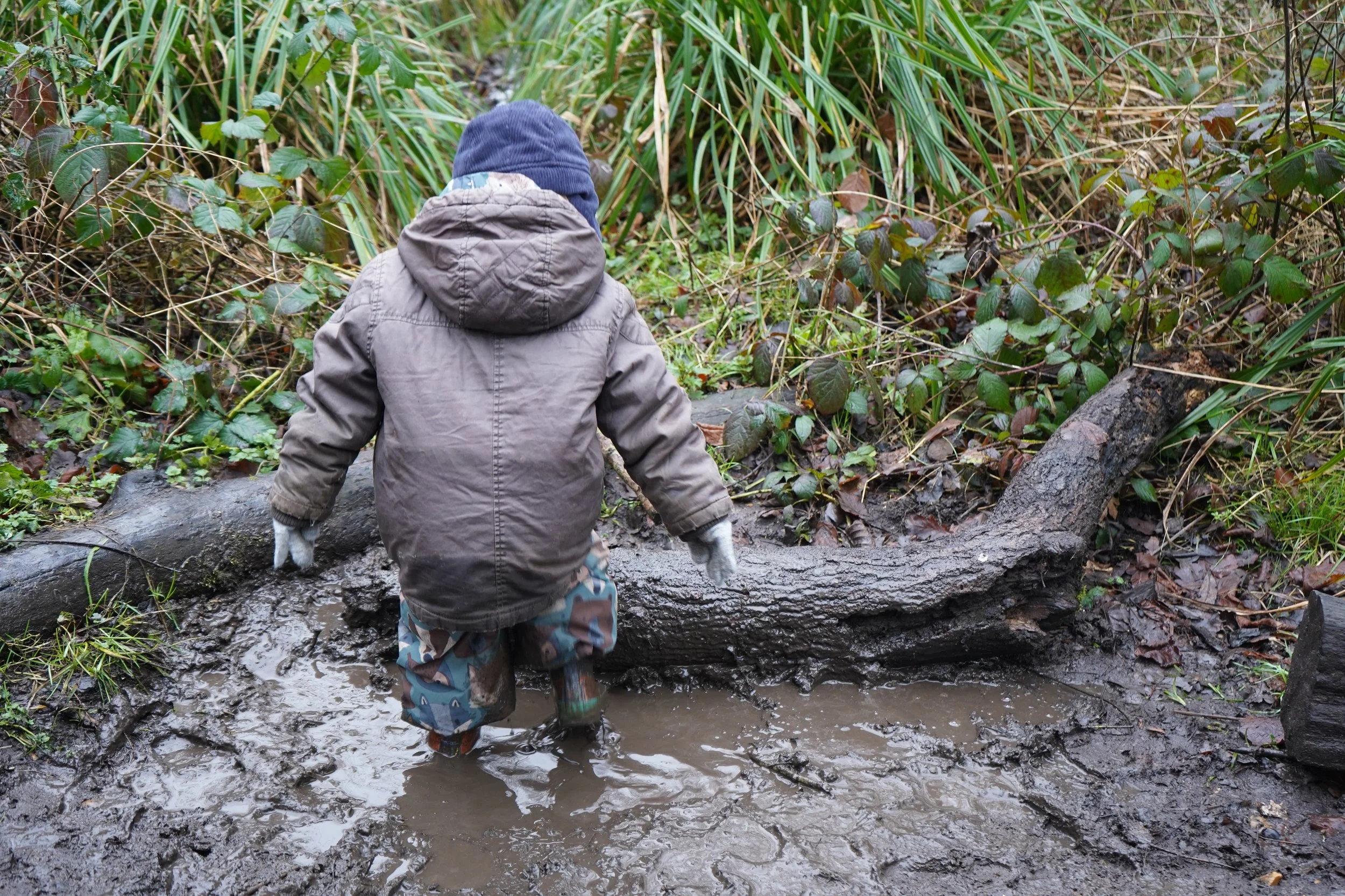 Child in a brown jacket and beanie walking through muddy puddles in a wooded area in Woodham Walter. 