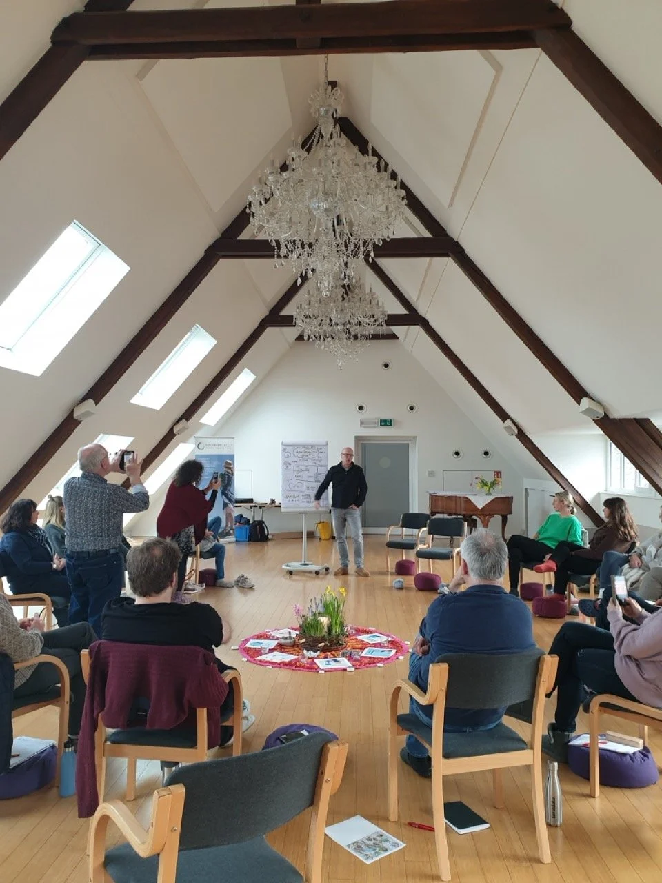 Menschen nehmen an einem Seminar oder Workshop in einem hellen Raum mit Holzboden und einem Kronleuchter teil, einige fotografieren, andere hören zu.