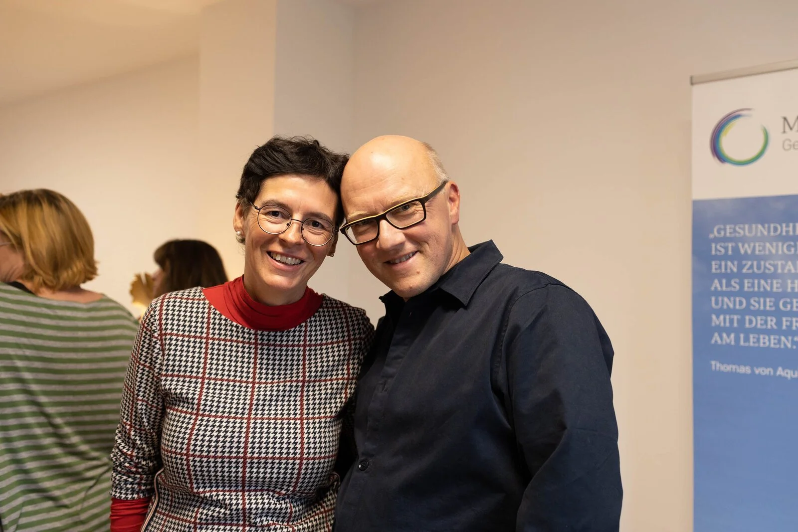 Zwei Menschen, eine Frau und ein Mann, die Seite an Seite stehen und in die Kamera lächeln, während sie bei einer Veranstaltung sind.