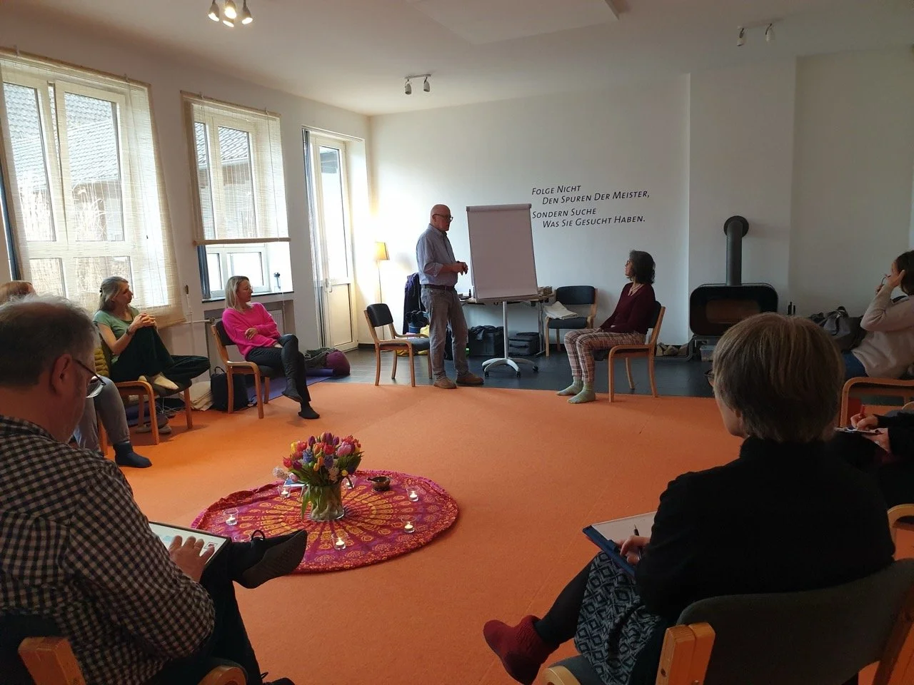 Gruppentherapiesitzung mit einem positiven, entspannten Ambiente, eine Person steht an einem Flipchart, während mehrere Teilnehmer auf Stühlen sitzen und zuhören, im Raum ist ein Kaminofen und eine bunte Dekoration auf einem Teppich mit Blumen.