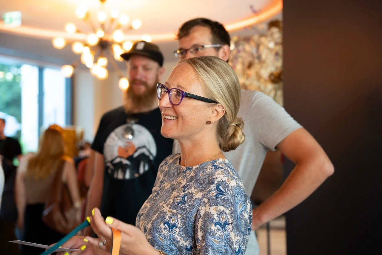 Drei Menschen in einem Raum, die Frau im Vordergrund trägt eine Brille und lächelt, während sie eine Zeitung hält. Die zwei Männer im Hintergrund sind ebenfalls lächelnd, der Raum ist modern gestaltet mit einer großen Lichtleiste an der Decke.