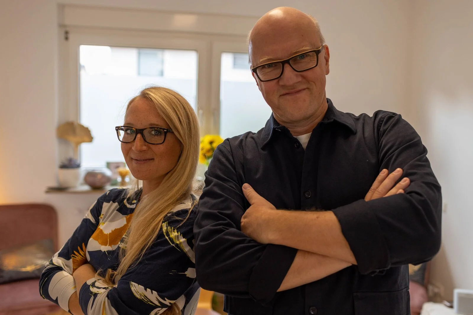 Ein Mann und eine Frau stehen nebeneinander in einem Wohnzimmer, beide mit verschränkten Armen und lächelnd. Der Mann trägt eine schwarze Hemd und trägt Brille, die Frau trägt ein Muster Kleid und trägt ebenfalls Brille.