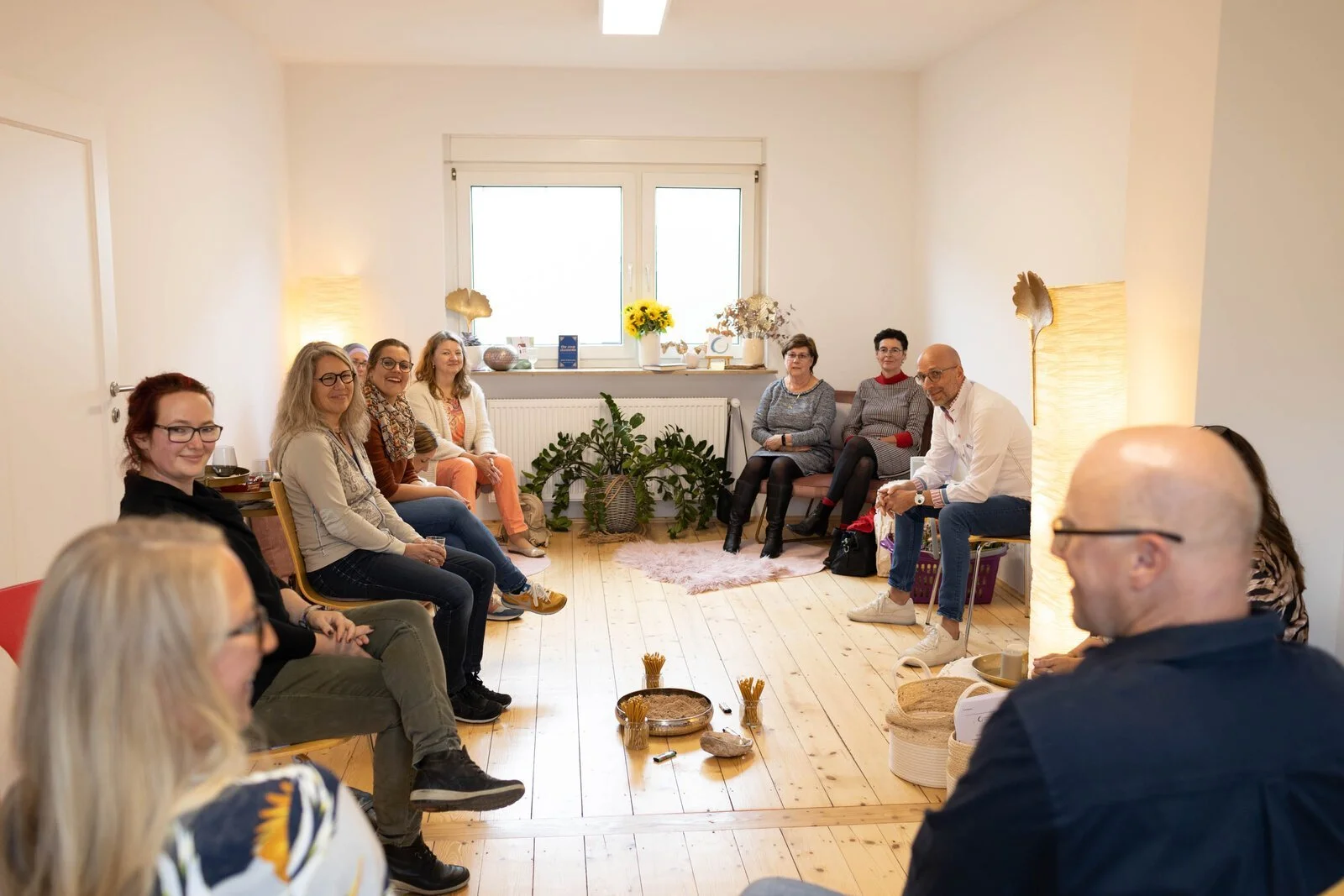 Menschen sitzen in einem ruhigen Raum im Kreis, der Raum ist hell erleuchtet mit natürlichem Licht durch das Fenster, dekoriert mit Pflanzen, Kerzen und Blumen. Sie scheinen in einem Workshop oder einer Gruppenaktivität zu sein.