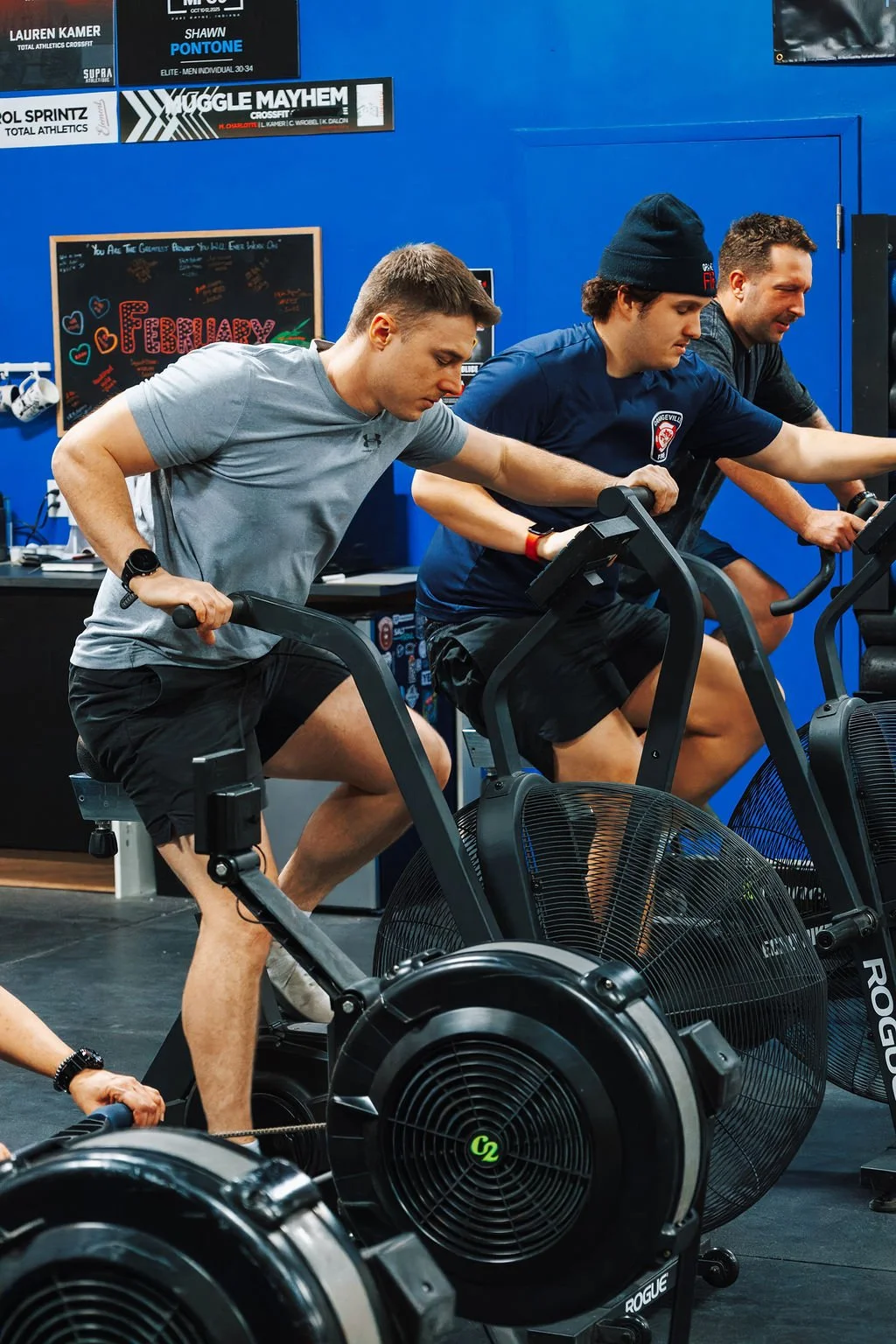 Three men using rowing machines during a workout session in a blue-walled gym with posters and a chalkboard in the background.