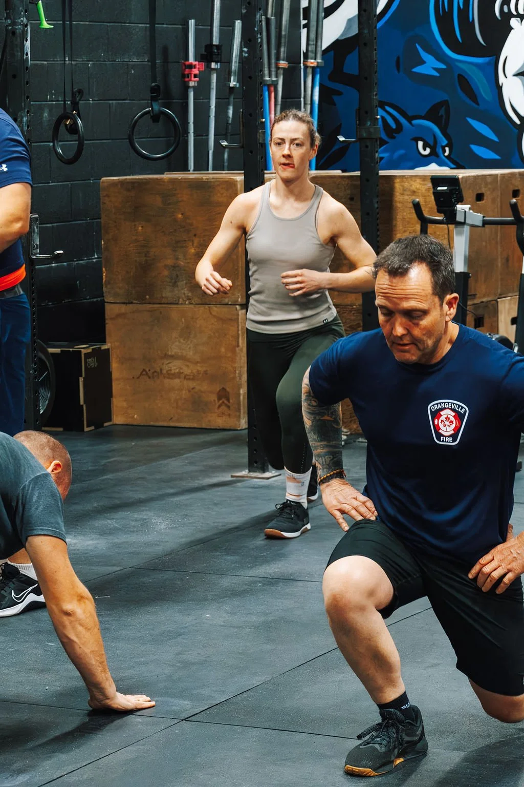 People exercising in a gym, with a man in a blue shirt and black shorts doing lunges, a woman in a gray tank top and black leggings running in place, and another person in a gray t-shirt and dark pants stretching or on the floor.