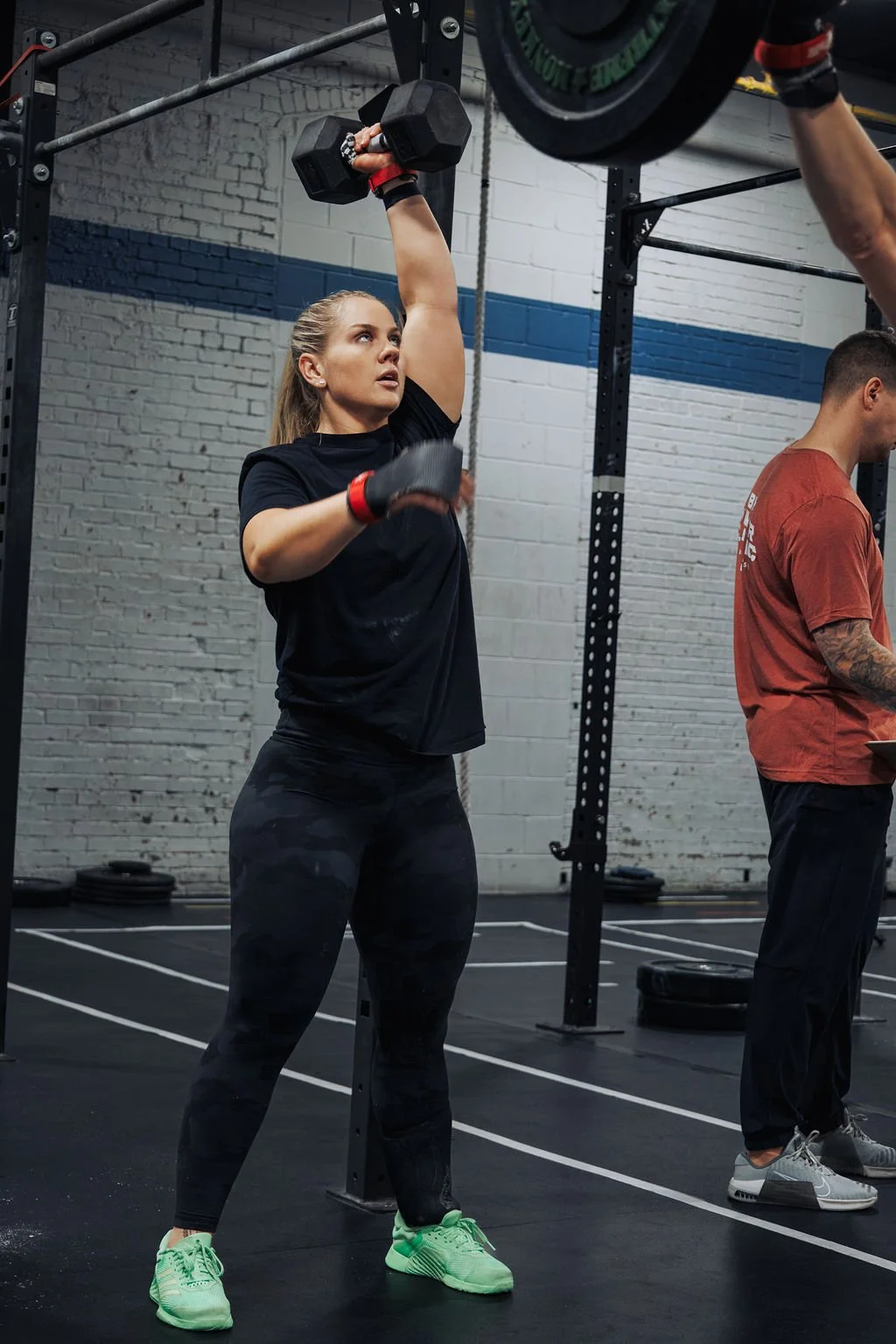 A woman in black athletic wear lifting a dumbbell overhead in a gym, with a focused expression. Another person stands nearby, looking downward. The gym has white brick walls with blue stripe accents.