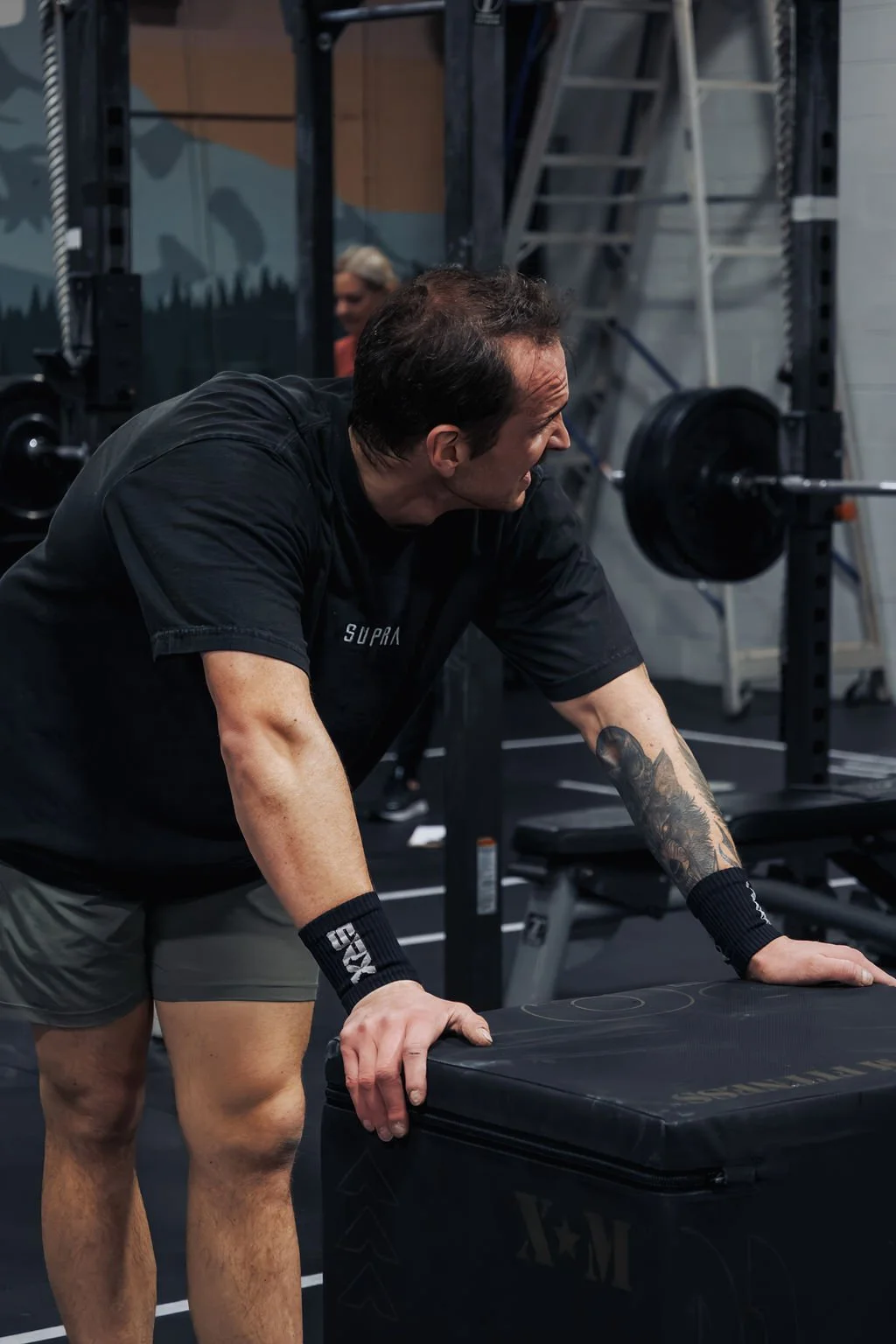 A man wearing a black T-shirt and gray shorts is exercising in a gym, leaning on a black padded box used for workouts, with gym equipment like a barbell and a weighing rack visible in the background.