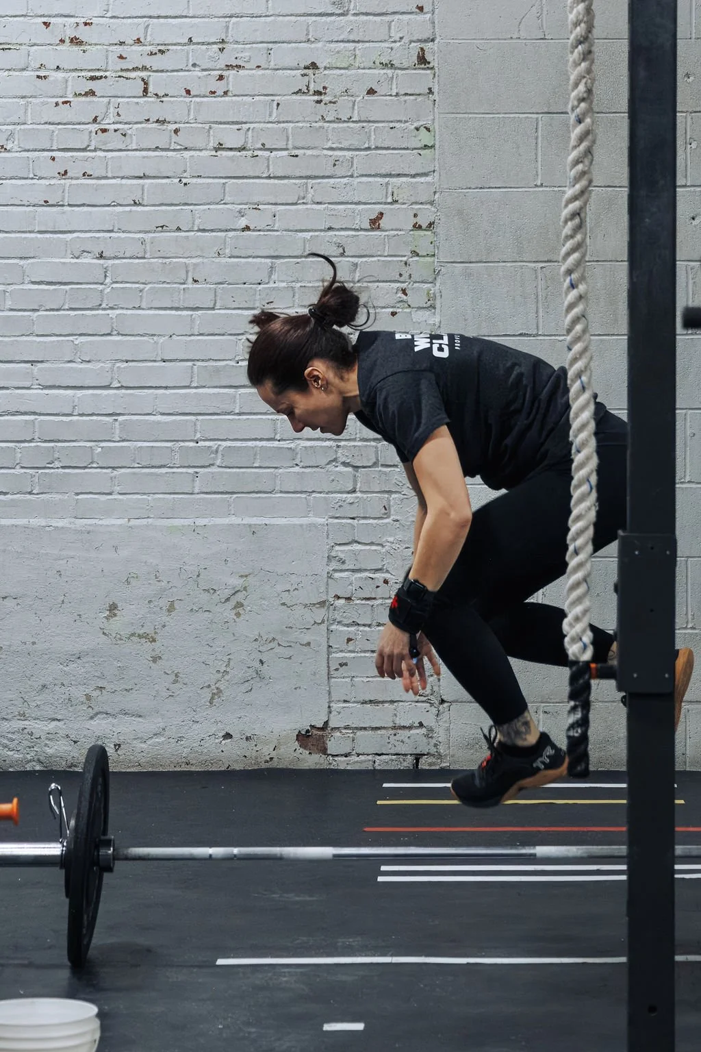 A woman performing a jump in a gym with a barbell on the ground, wearing black workout clothes and wristbands, against a white brick wall.