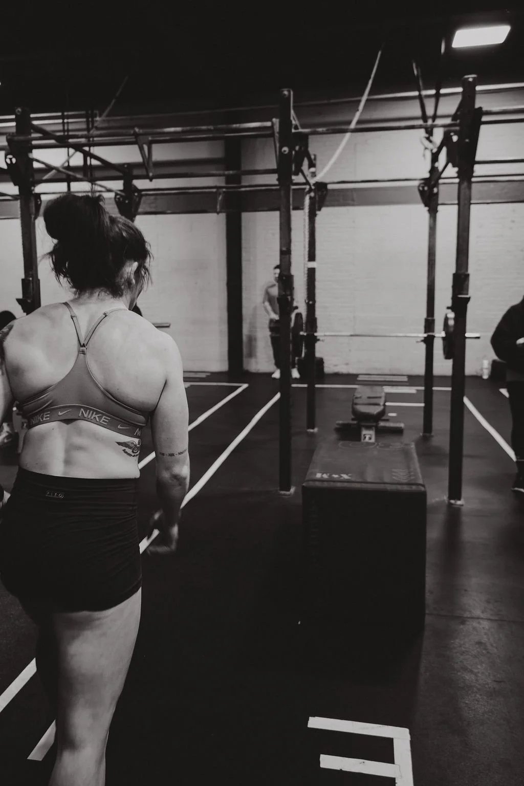 A woman in a sports bra and shorts standing in a gym near a plyometric box and a pull-up bar, with other people in the background.