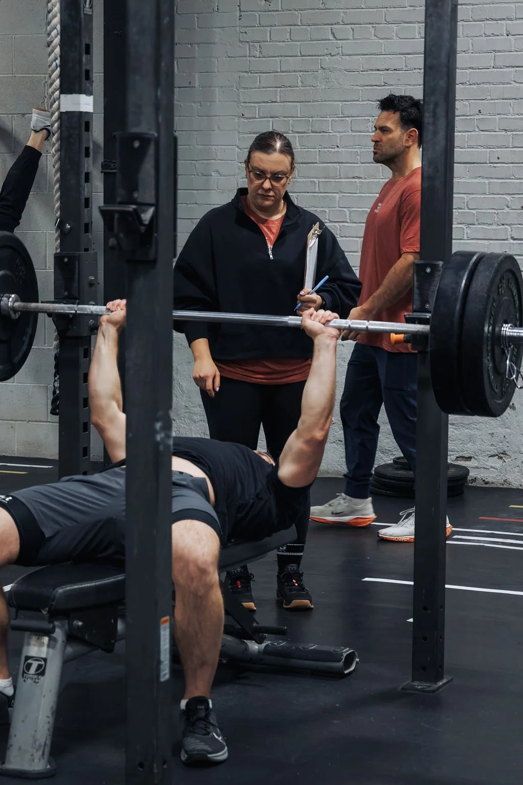 A person lying on a bench press, lifting a barbell with weights while two trainers observe and take notes in a gym with gray brick walls.