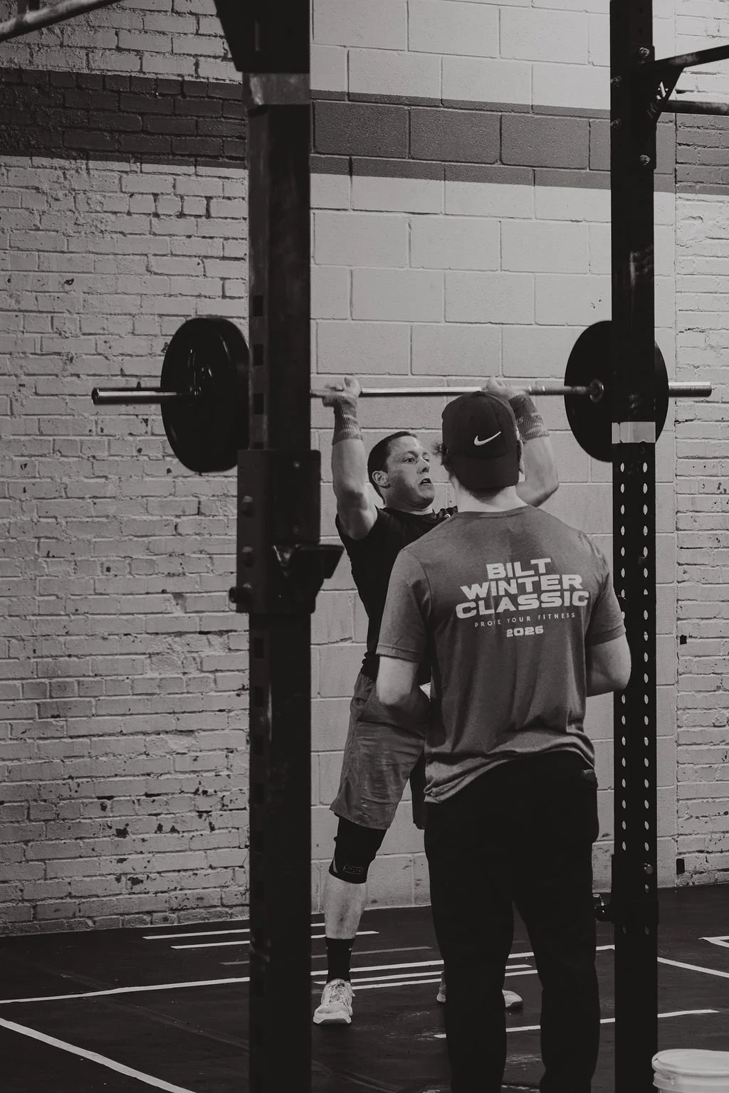 A man is lifting a barbell above his head during a workout session in a gym, with a trainer standing in front of him, wearing a t-shirt that reads "Bilt Winter Classic". The scene is in black and white, set against a brick wall background.