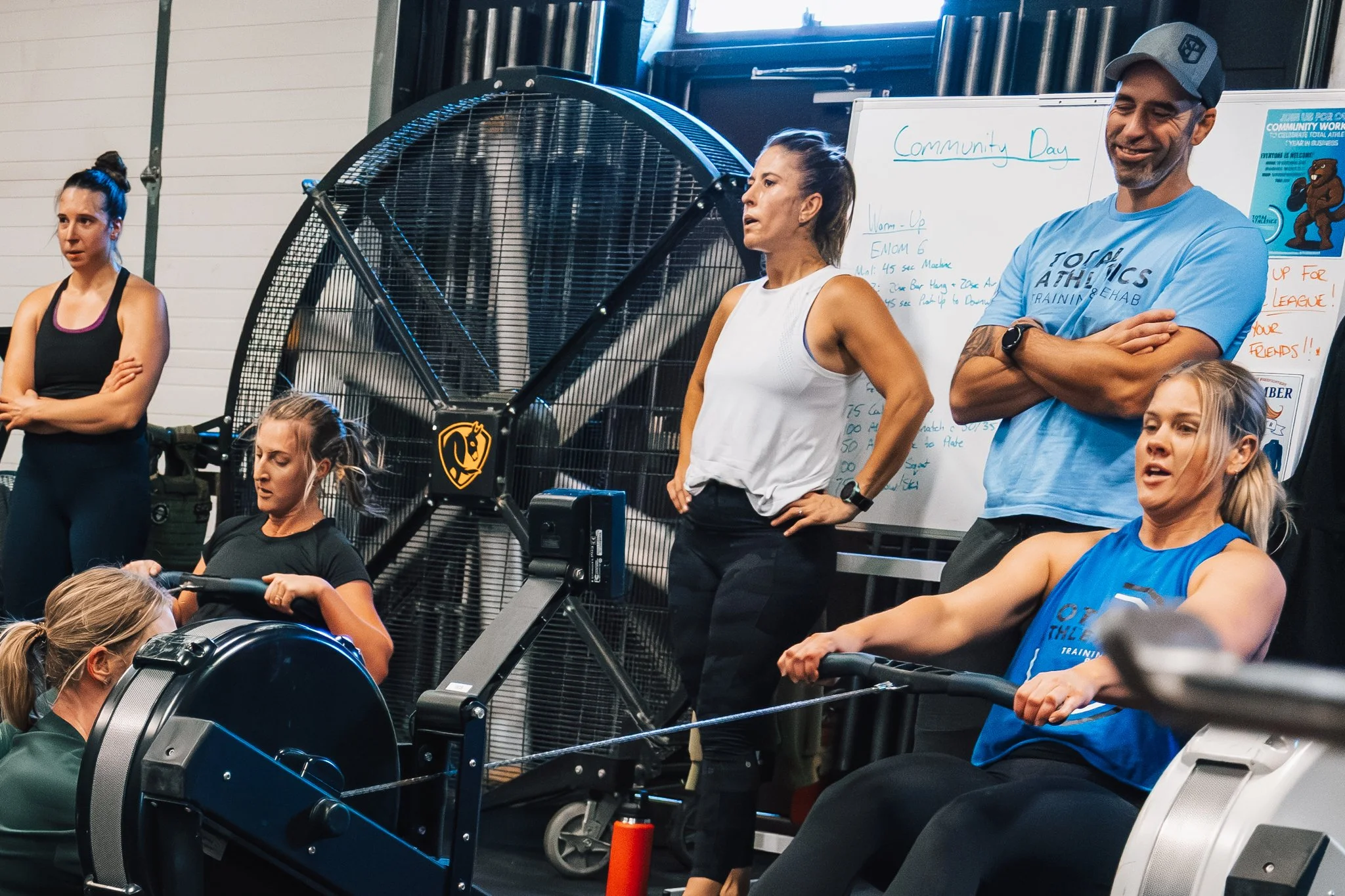 Group of people in a gym during a rowing workout class, with some participants rowing on machines and others standing and watching in front of a whiteboard that says 'Community Day'.