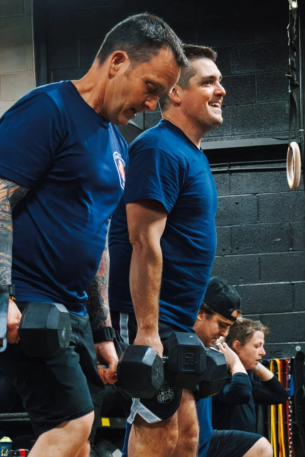 Two men lifting dumbbells during a workout in a gym, with others exercising in the background.